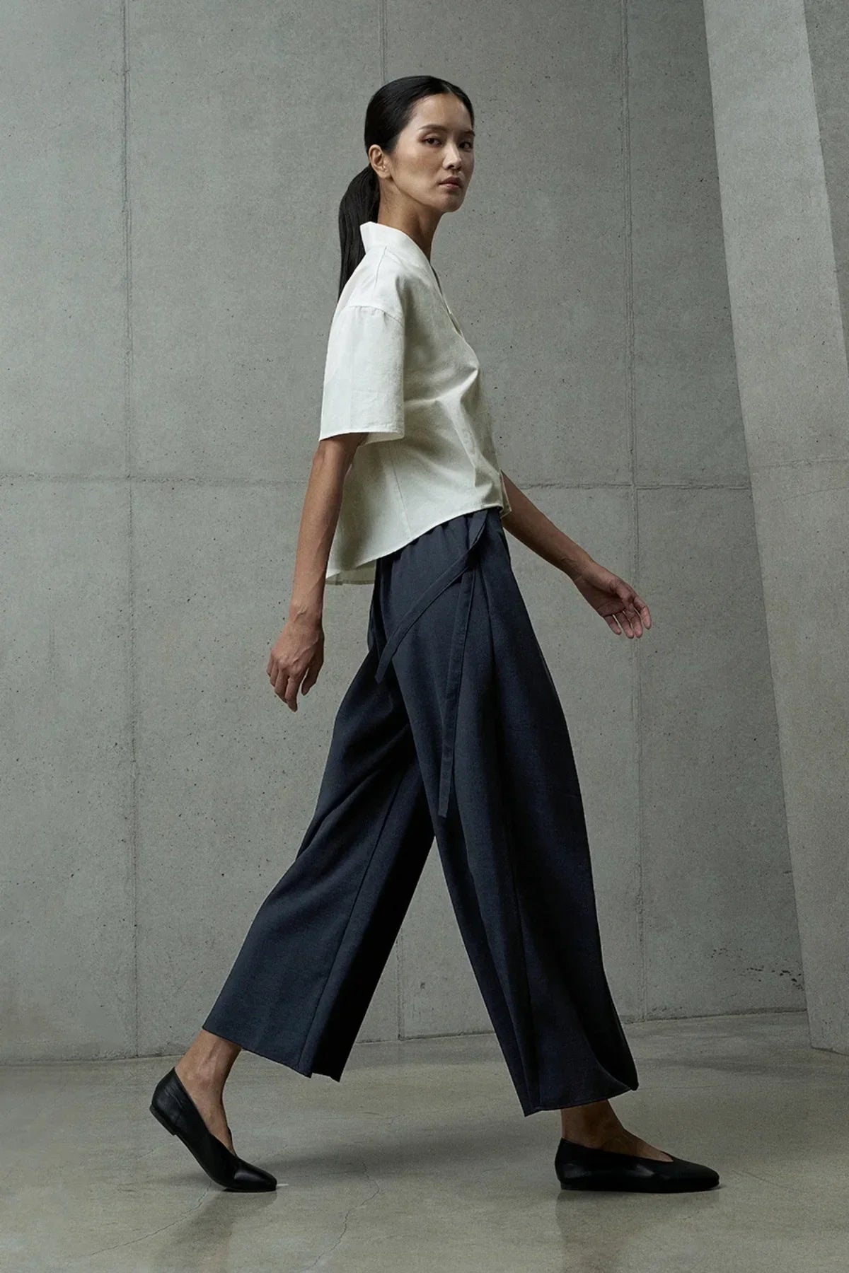 Woman walking in ivory short-sleeved shirt, wide-leg black pants, and black flats against gray concrete wall