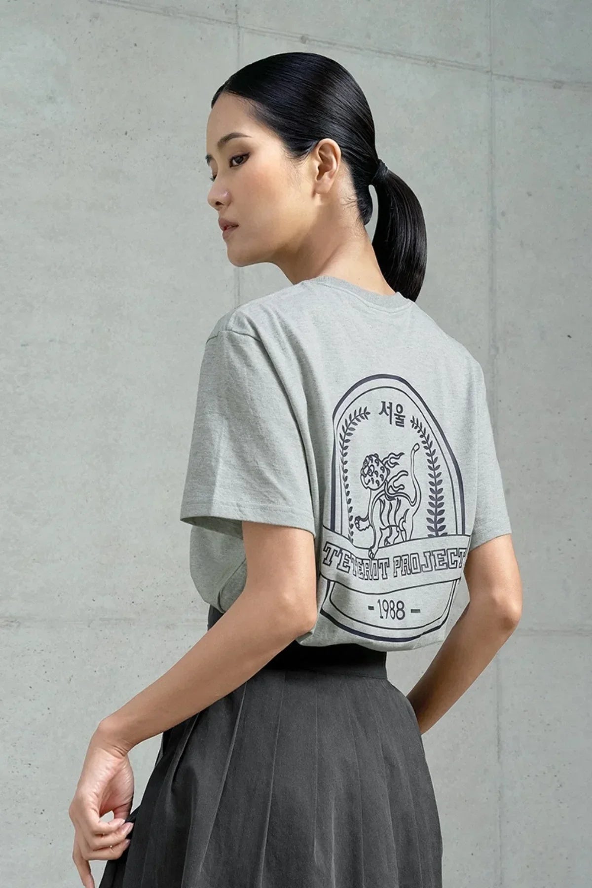 Woman wearing gray Seoul-themed T-shirt with tiger graphic and text, paired with dark pleated skirt