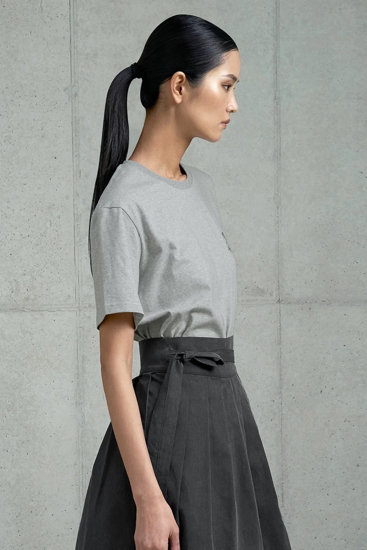 Side view of woman with long ponytail wearing a gray t-shirt and high-waisted pleated black skirt against concrete background