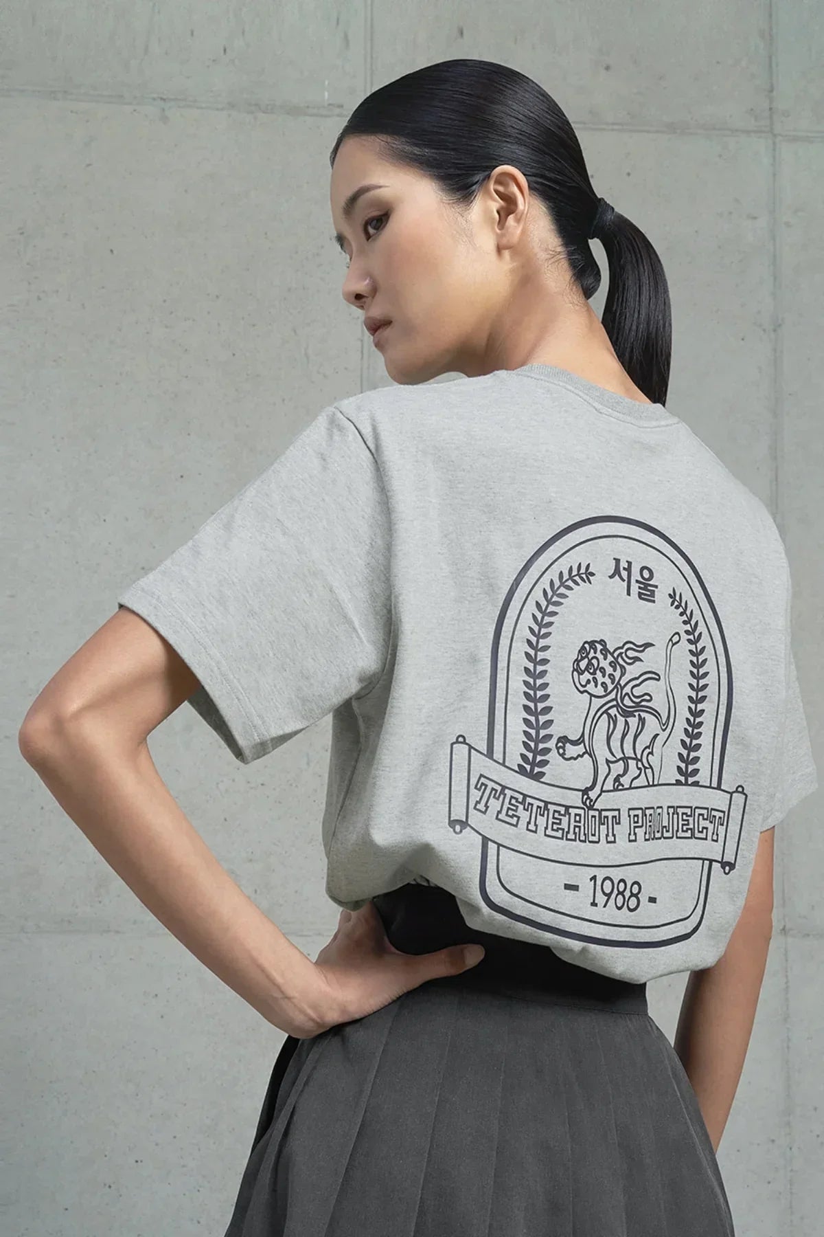 Woman wearing gray T-shirt with Seoul text and tiger graphic, posing against concrete wall