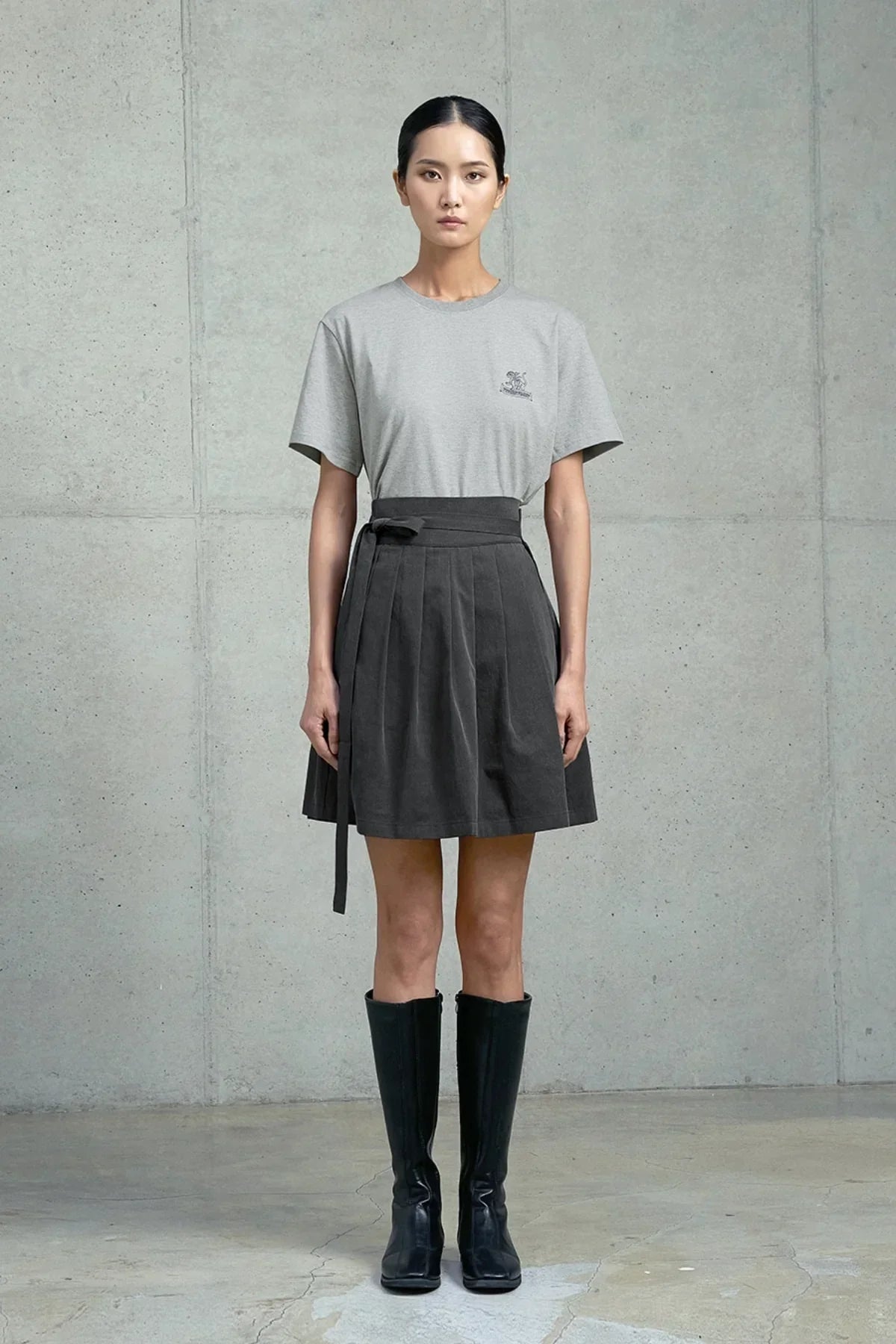 Woman wearing gray Hachi Seoul t-shirt with logo, dark pleated skirt and black boots against concrete wall