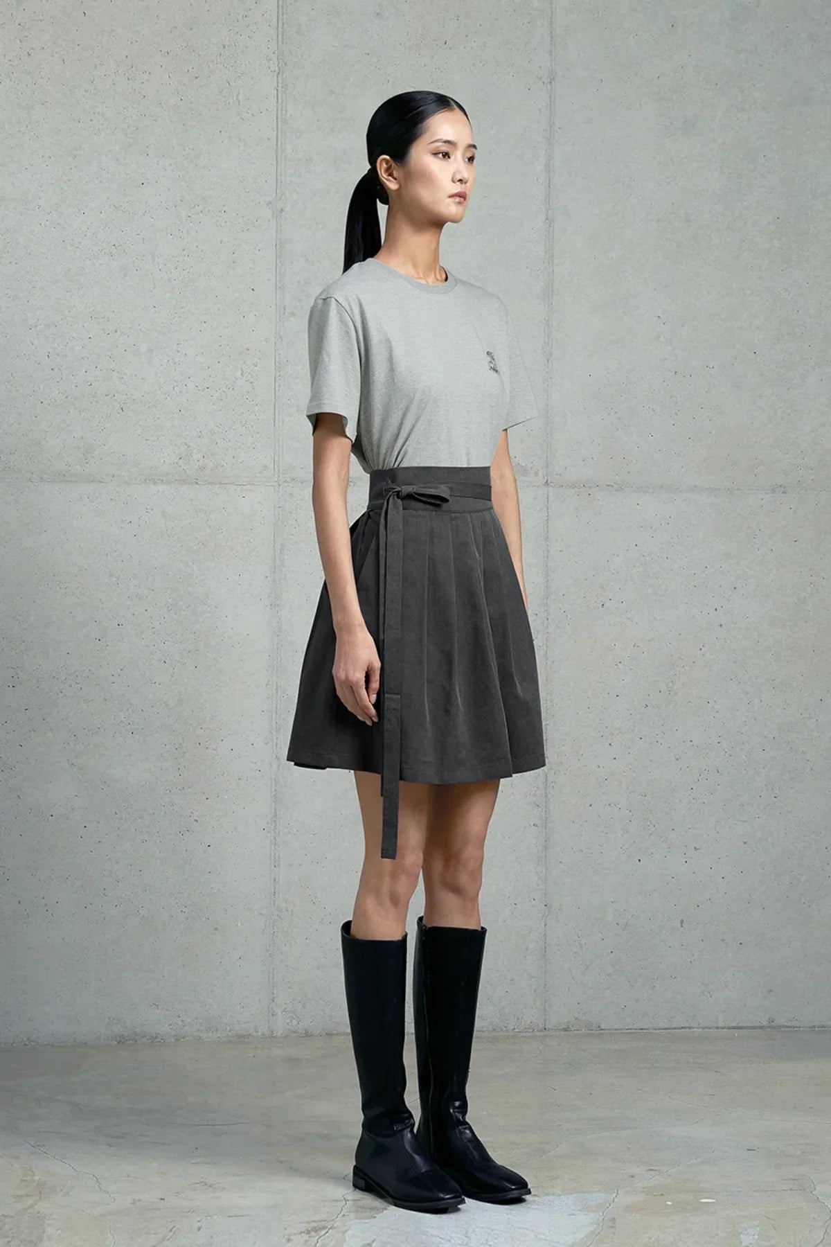 Model wearing gray graphic t-shirt, dark pleated skirt with belt, and black knee-high boots against concrete wall