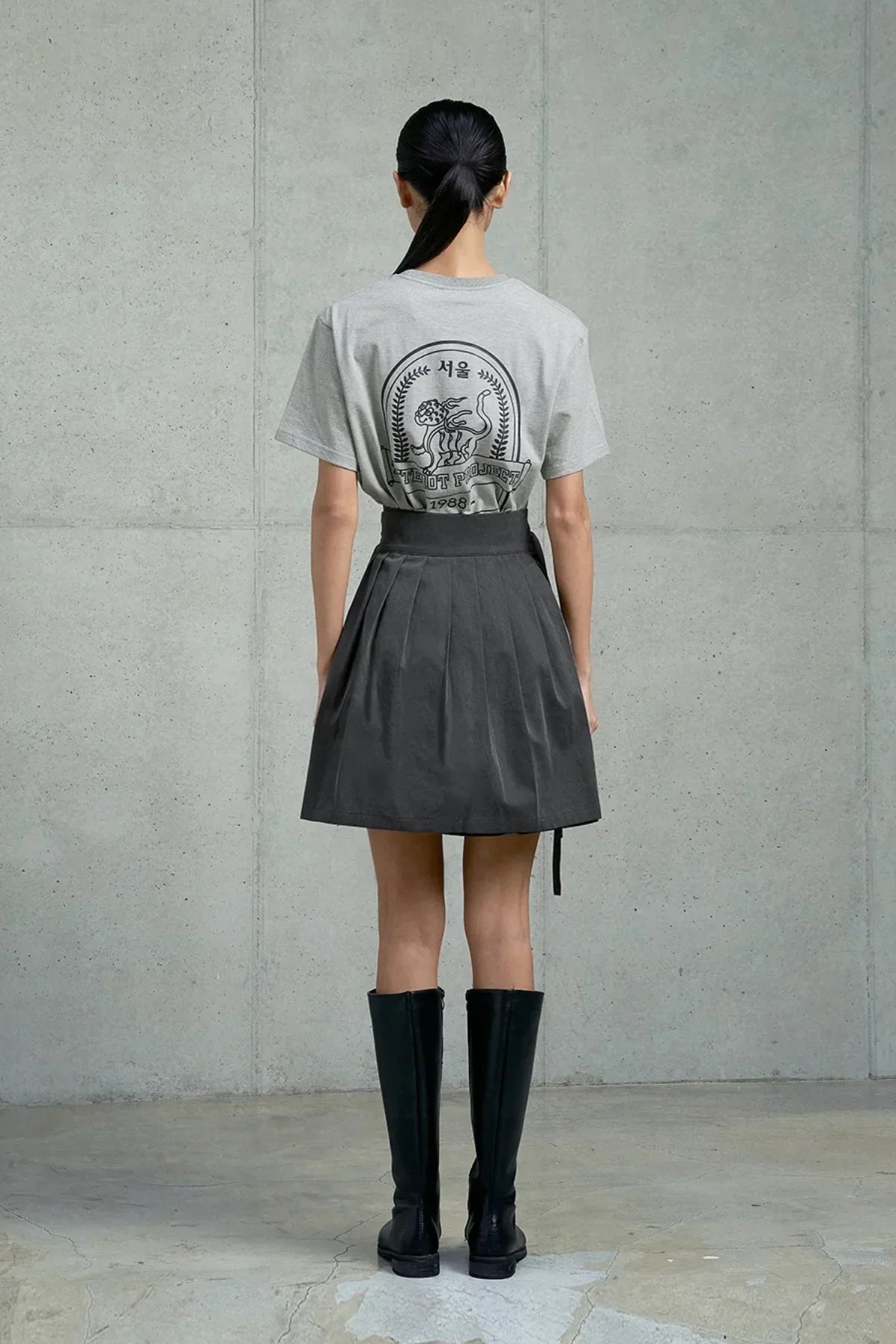 Model wearing gray Seoul graphic t-shirt, black pleated skirt, and knee-high black boots, standing indoors