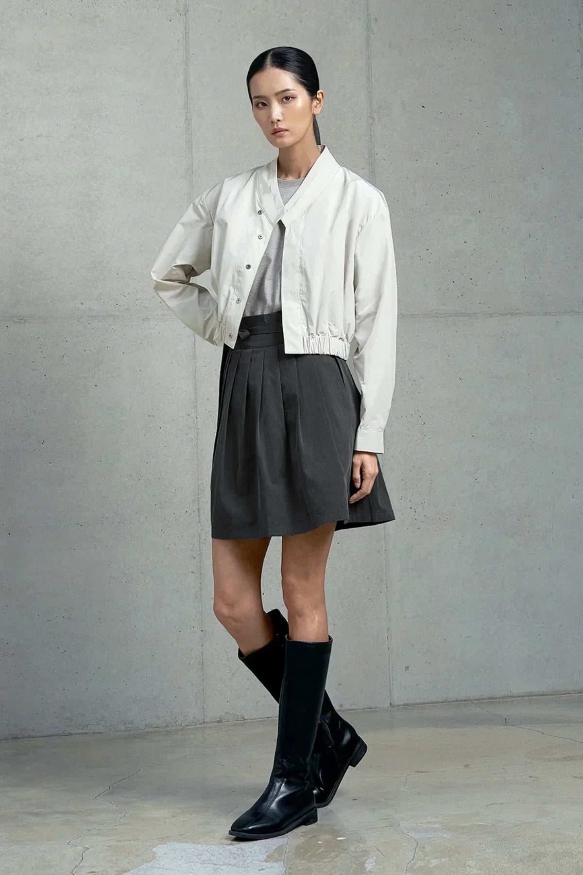 Model wearing gray t-shirt, white cropped jacket, black pleated skirt, and black knee-high boots in studio