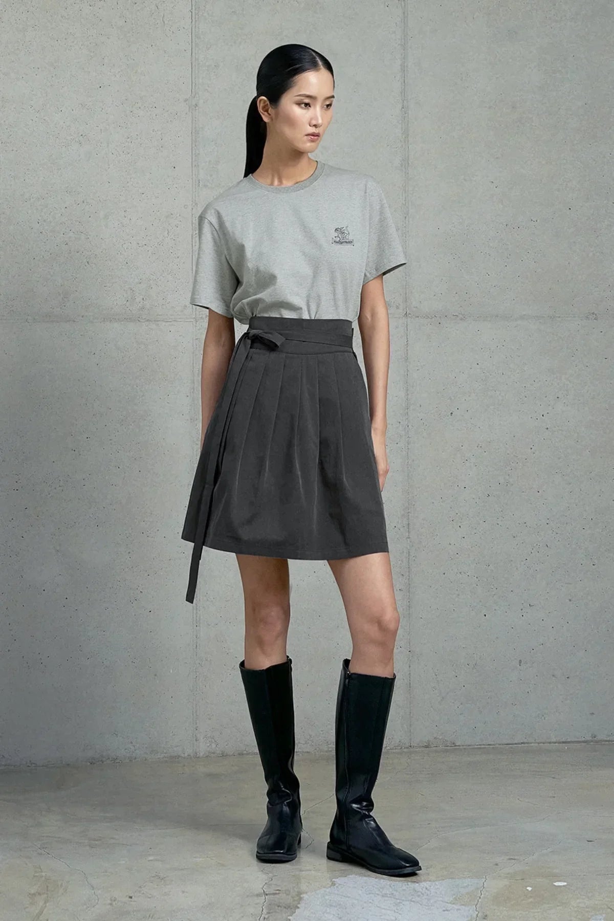 Woman wearing gray graphic t-shirt, black pleated skirt, and knee-high black boots against concrete wall