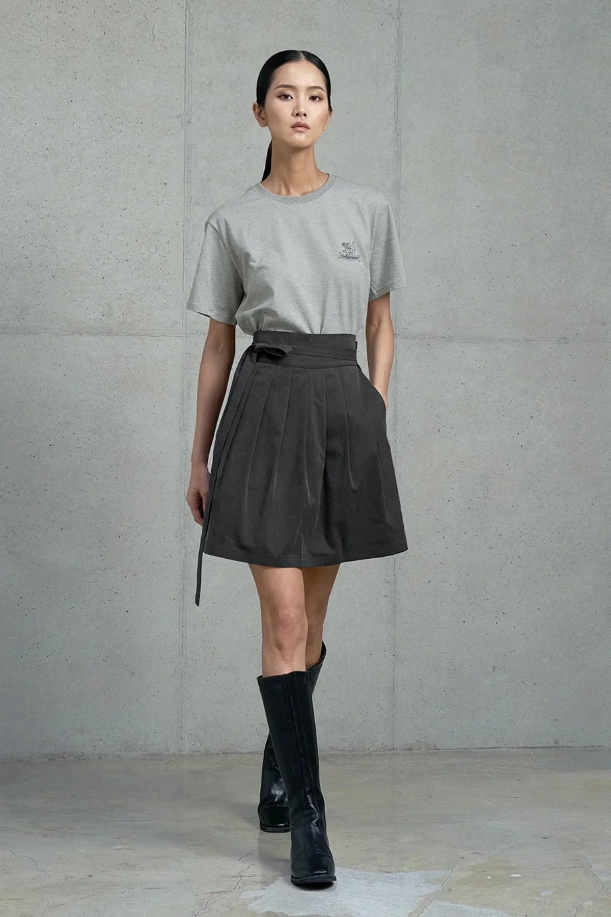 Woman modeling gray graphic t-shirt with dark pleated skirt and black knee-high boots in studio