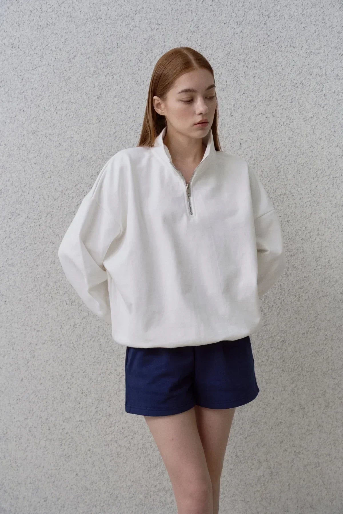 Model wearing white half-zip sweatshirt and navy shorts against textured gray wall