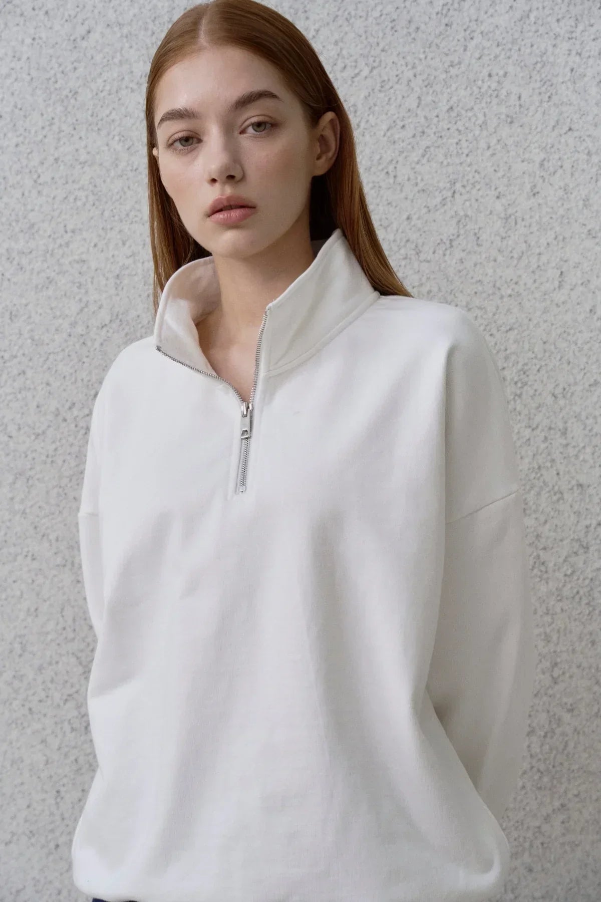 Young woman wearing a white half-zip sweatshirt against a textured gray wall