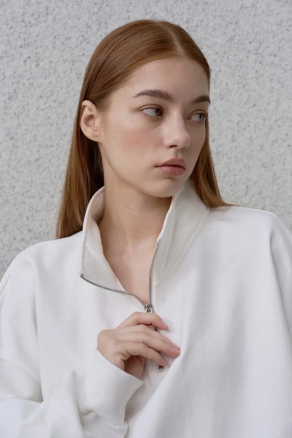 Young woman wearing white half-zip sweatshirt with high collar against grey wall