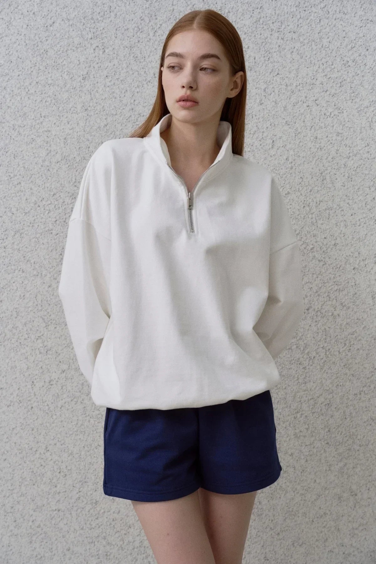 Young woman wearing a white half-zip sweatshirt and navy shorts against a textured gray wall