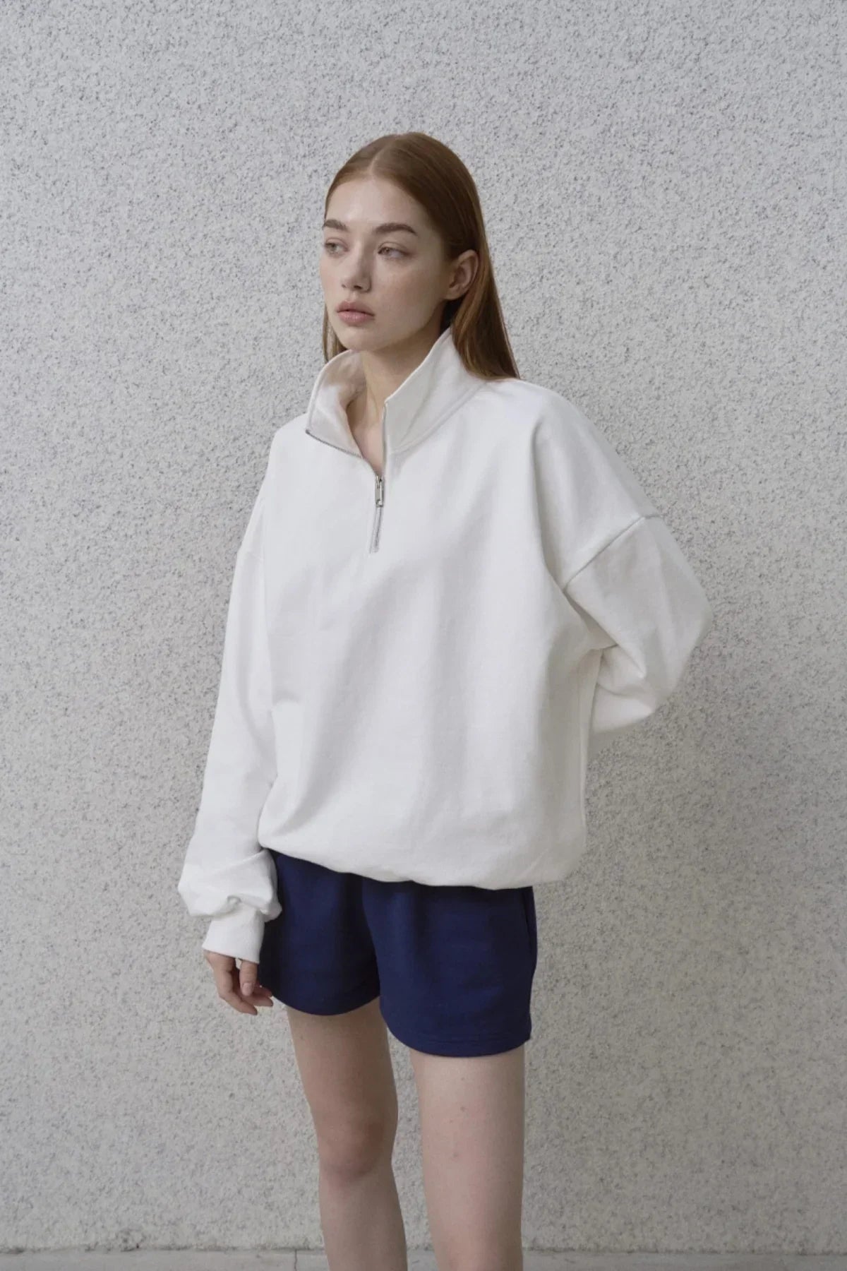 Woman wearing white half-zip sweatshirt and navy blue shorts standing against textured gray wall