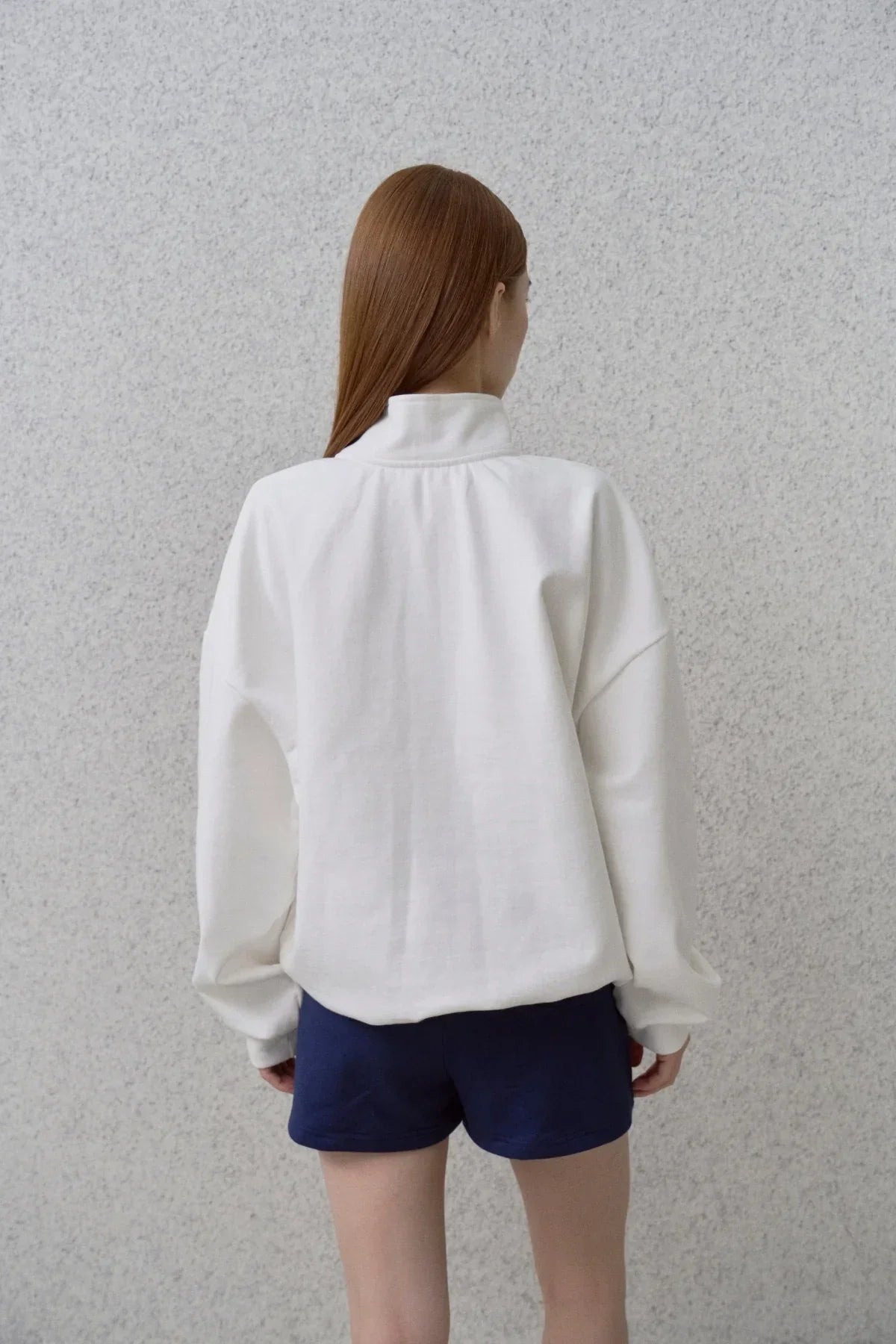 Woman wearing white half-zip sweatshirt and navy shorts standing against gray wall, back view