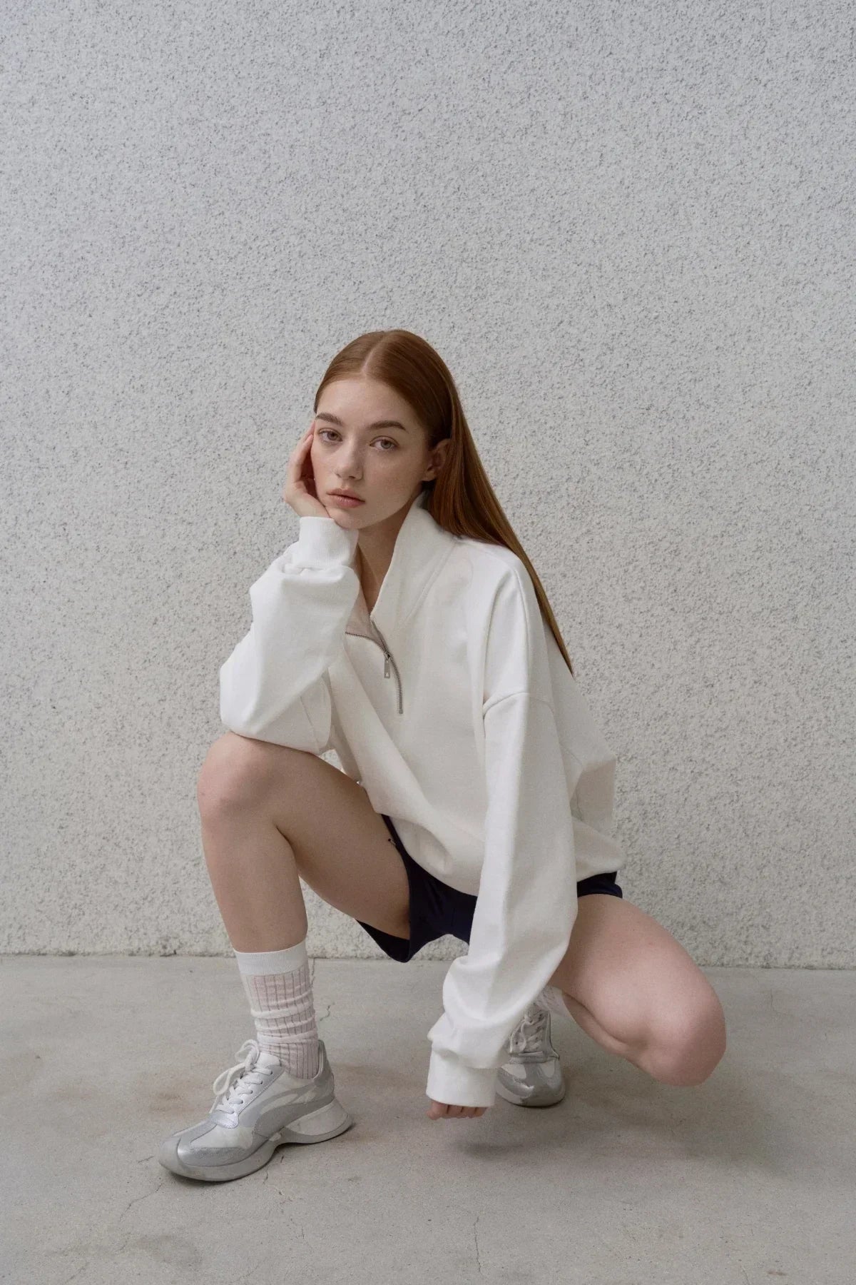 Young woman with red hair wearing white half-zip sweatshirt, black shorts, and silver sneakers squatting indoors