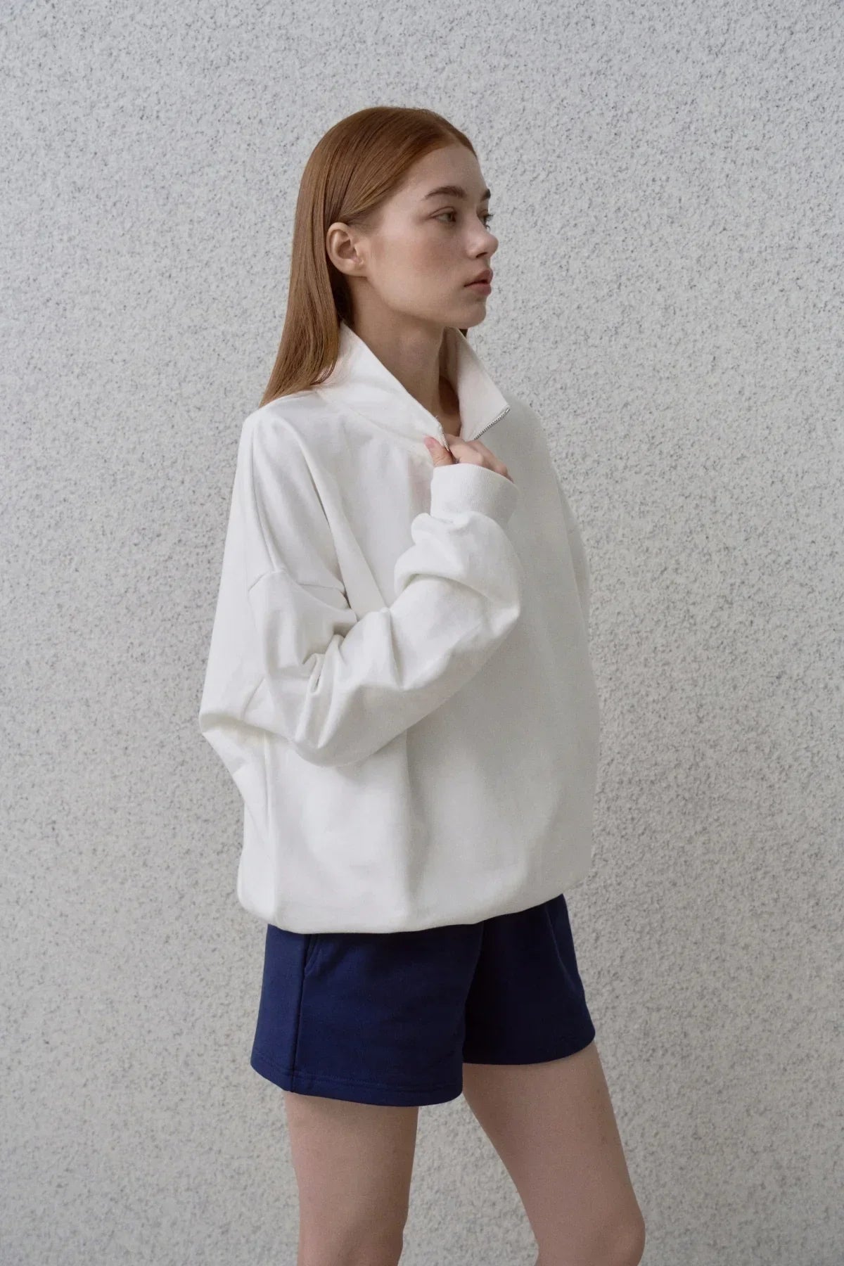 Side view of woman wearing white half-zip sweatshirt and navy shorts against gray textured wall