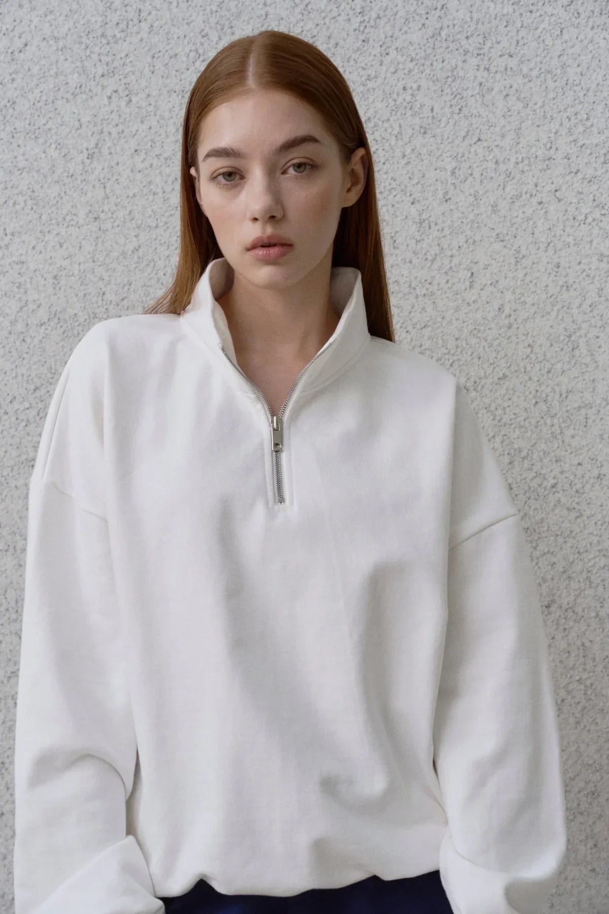 Young woman wearing a white half-zip sweatshirt standing against a textured gray wall