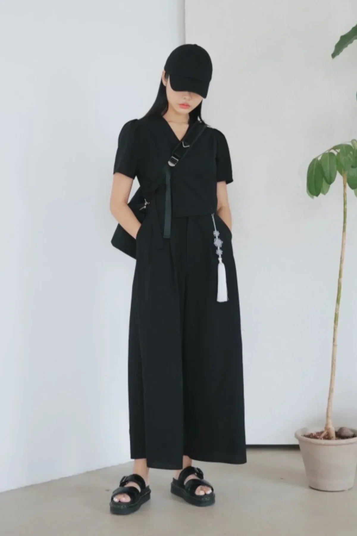 Woman wearing a black jumpsuit with short sleeves, black cap, and black sandals, standing indoors next to a potted plant