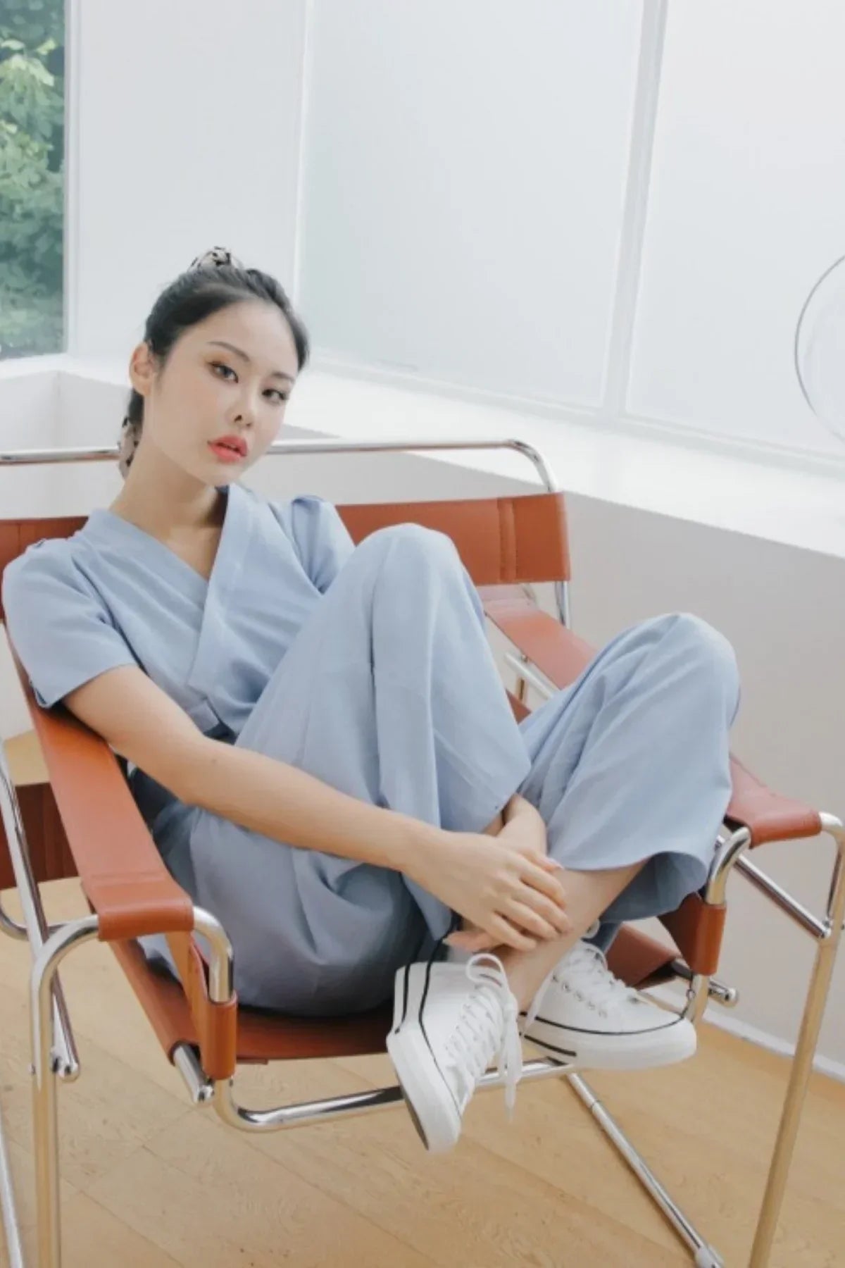 Woman in light blue jumpsuit sitting on modern brown leather chair with white sneakers