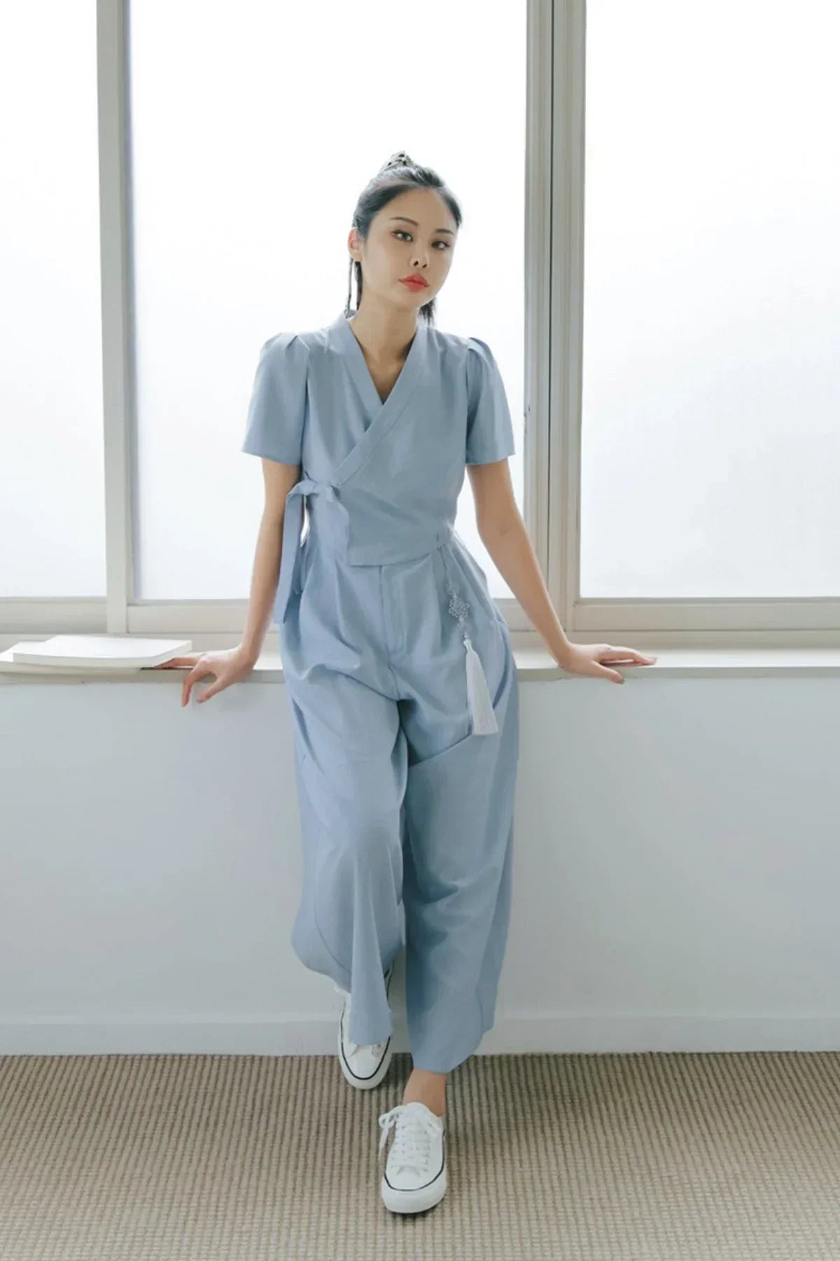 Woman wearing light blue wrap jumpsuit with short puff sleeves and white sneakers leaning on window sill indoors