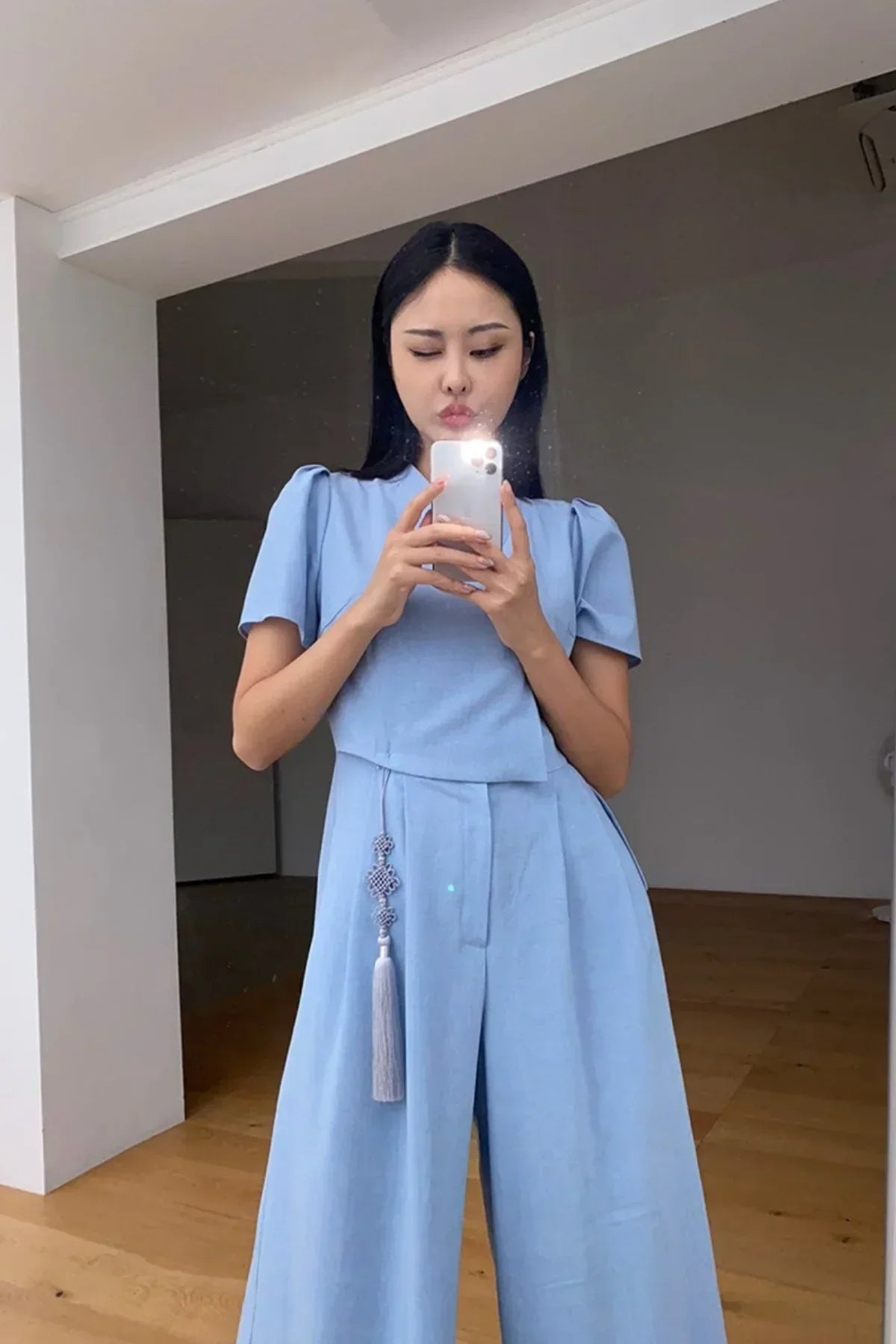 Woman wearing light blue jumpsuit with puff sleeves and tassel detail taking mirror selfie in minimalist room