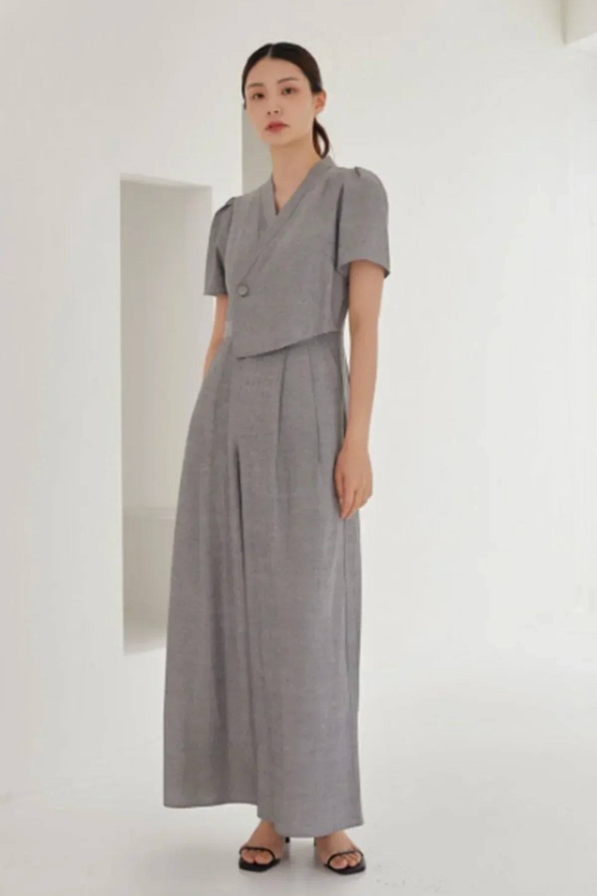 Woman wearing gray short-sleeve jumpsuit with wide-leg pants and black sandals in minimal white room