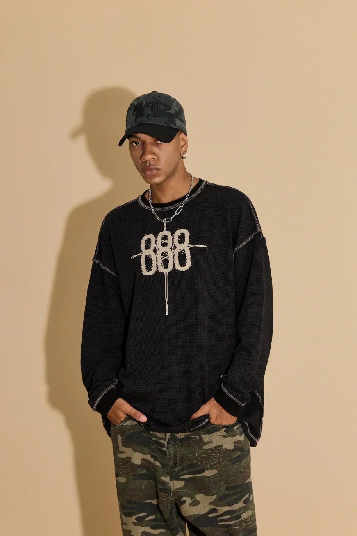 Man wearing black sweater with white graphic, camouflage pants, and grey camo ball cap against beige background