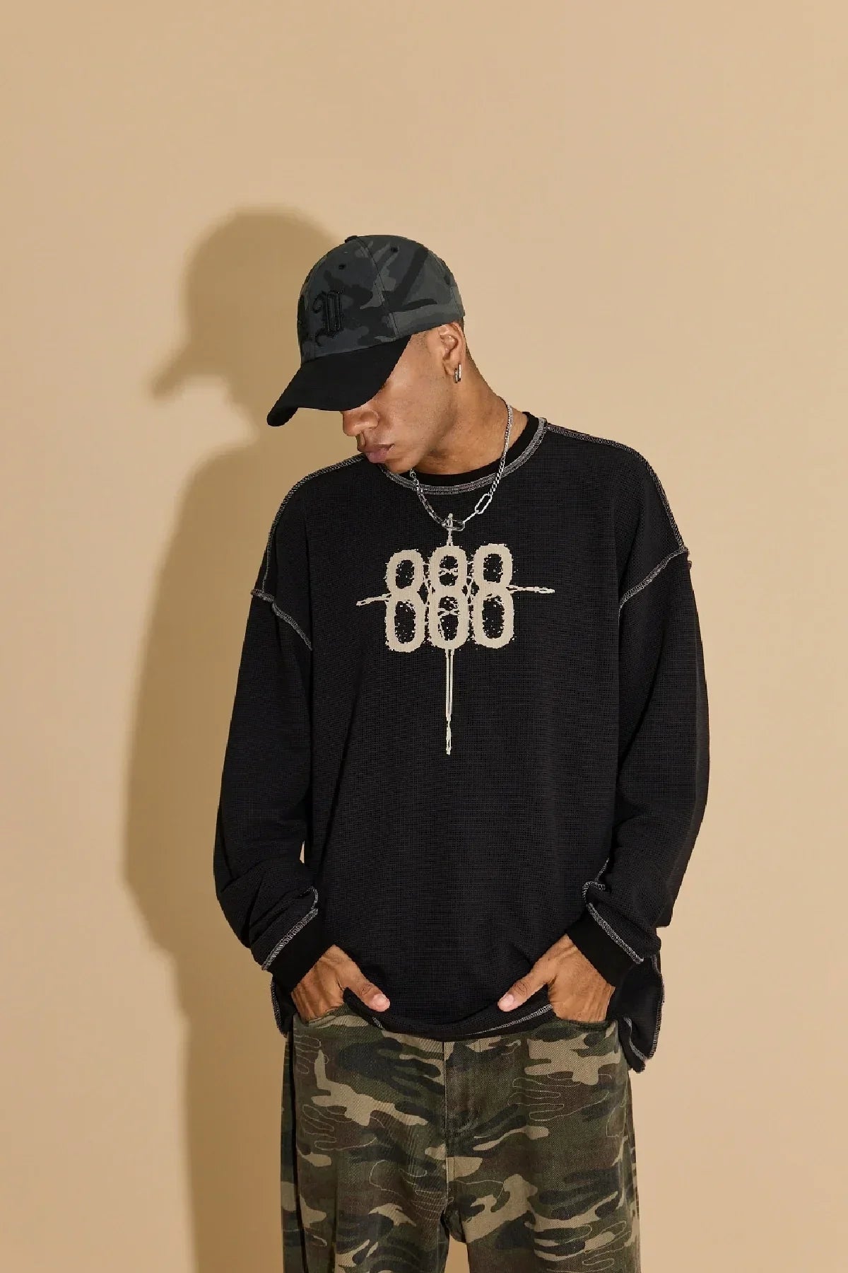 Man wearing black sweatshirt with 888 design, grey camo ball cap, and camo pants on beige background