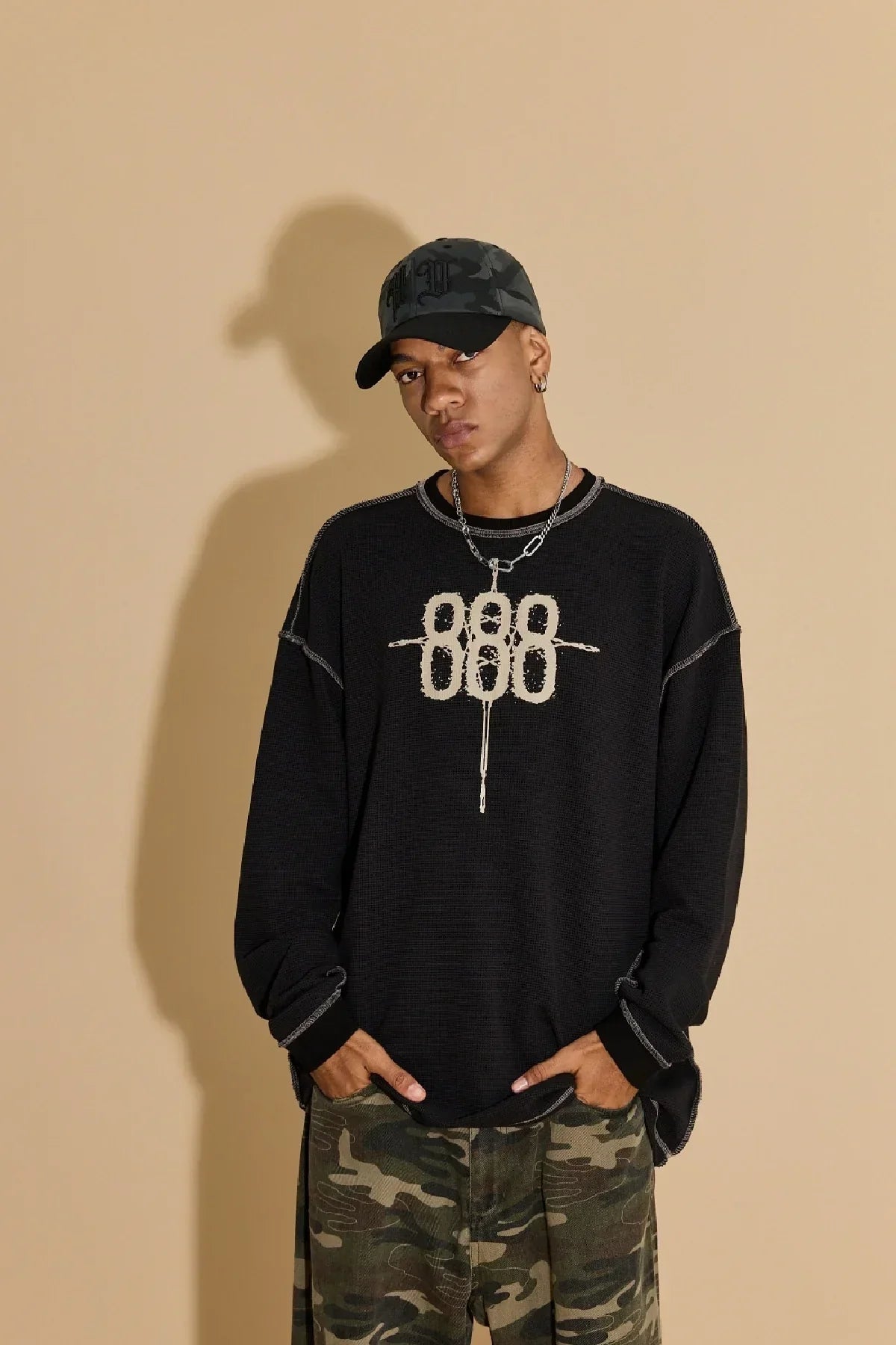 Man wearing black sweater with white 888 design, camo pants, and grey camo baseball cap against beige background