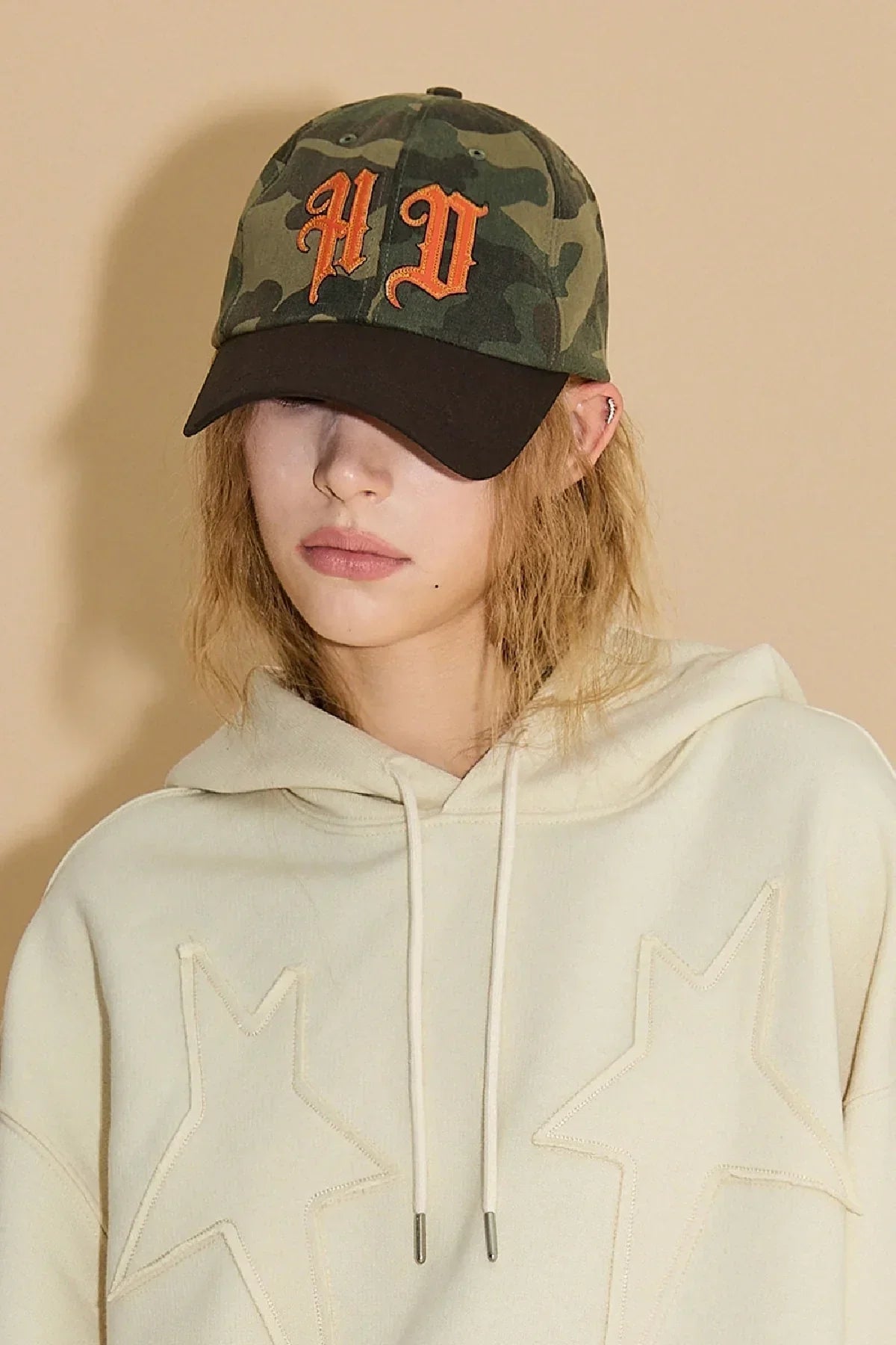 Close-up of a woman wearing a camo baseball cap with orange 'AD' letters and a beige hoodie with star patches