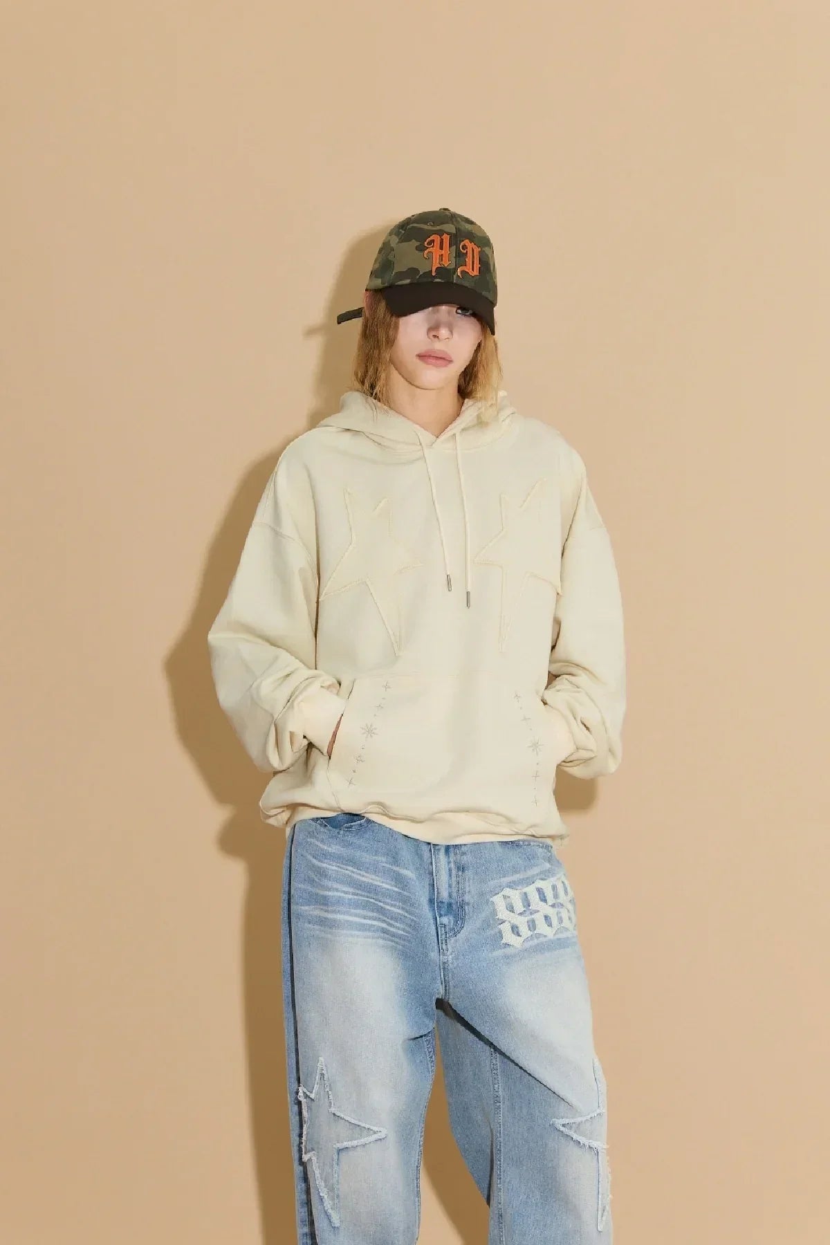 Person wearing beige hoodie with star design, light blue jeans with star patches, and camo baseball cap