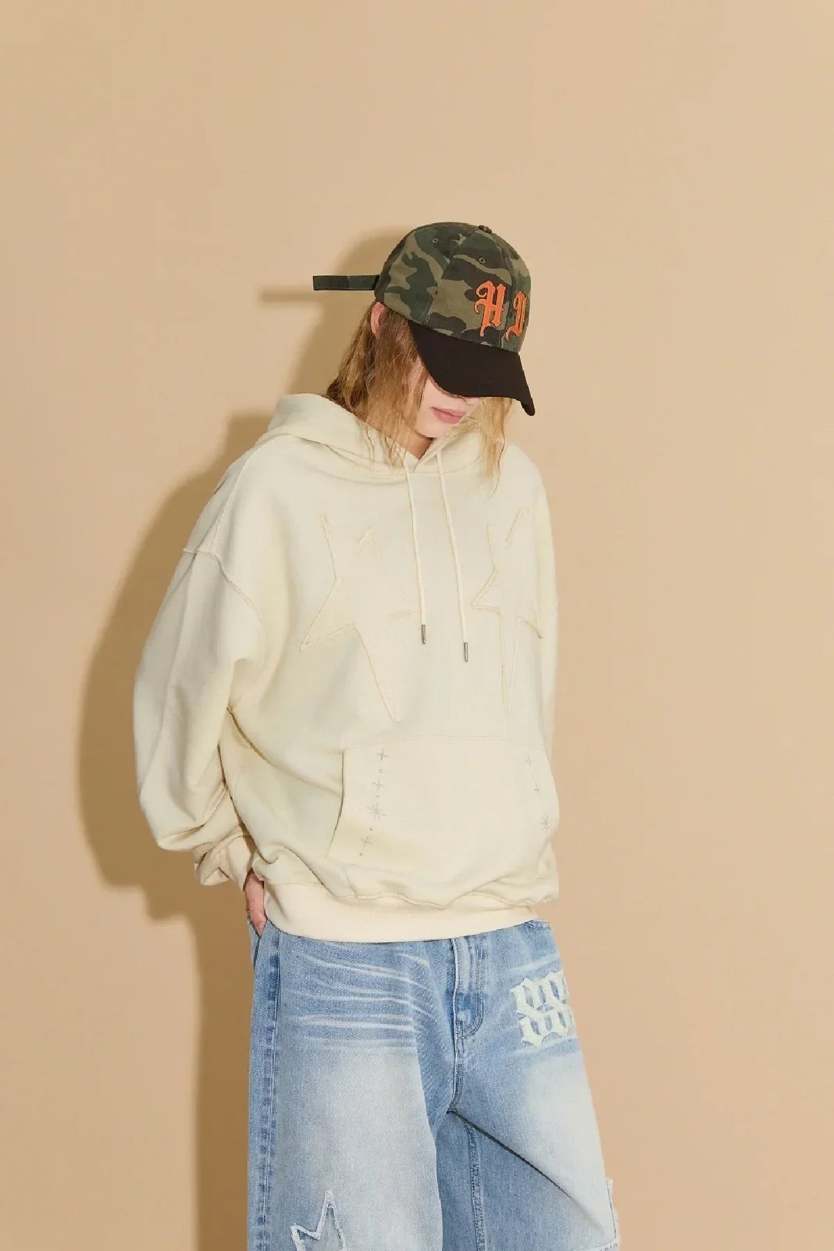 Person wearing a khaki camouflage ball cap with orange letters, cream hoodie with star appliques, and light blue jeans