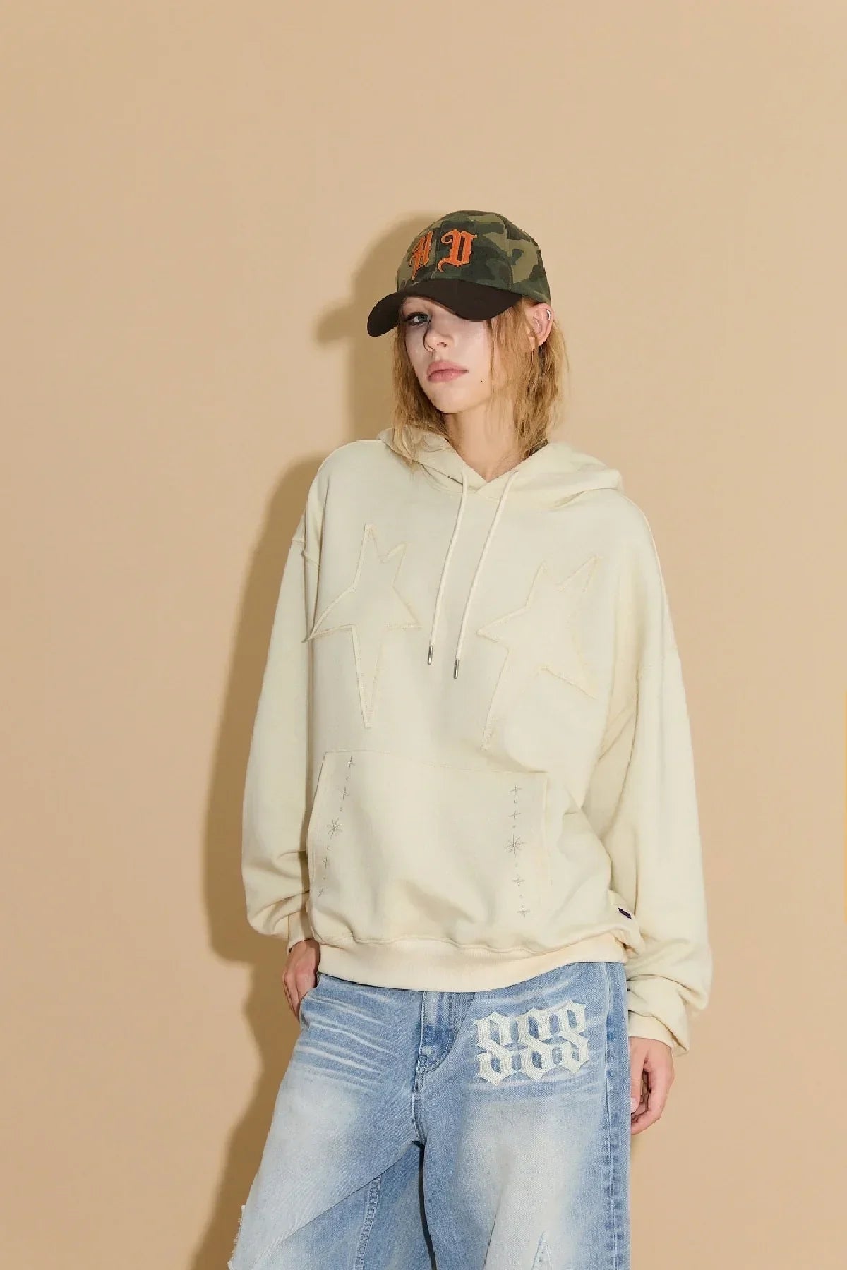 Model wearing beige star-appliqué hoodie, loose blue jeans with white design, and camo baseball cap with orange letters
