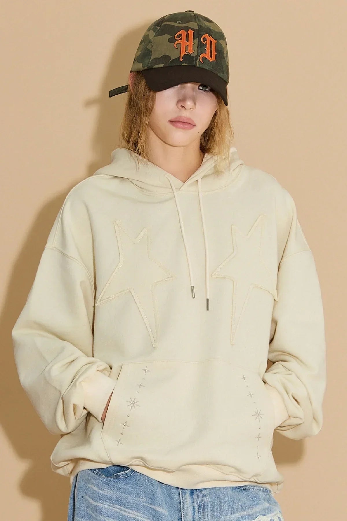Person wearing khaki camo ball cap with orange PD letters and beige hoodie with star appliques