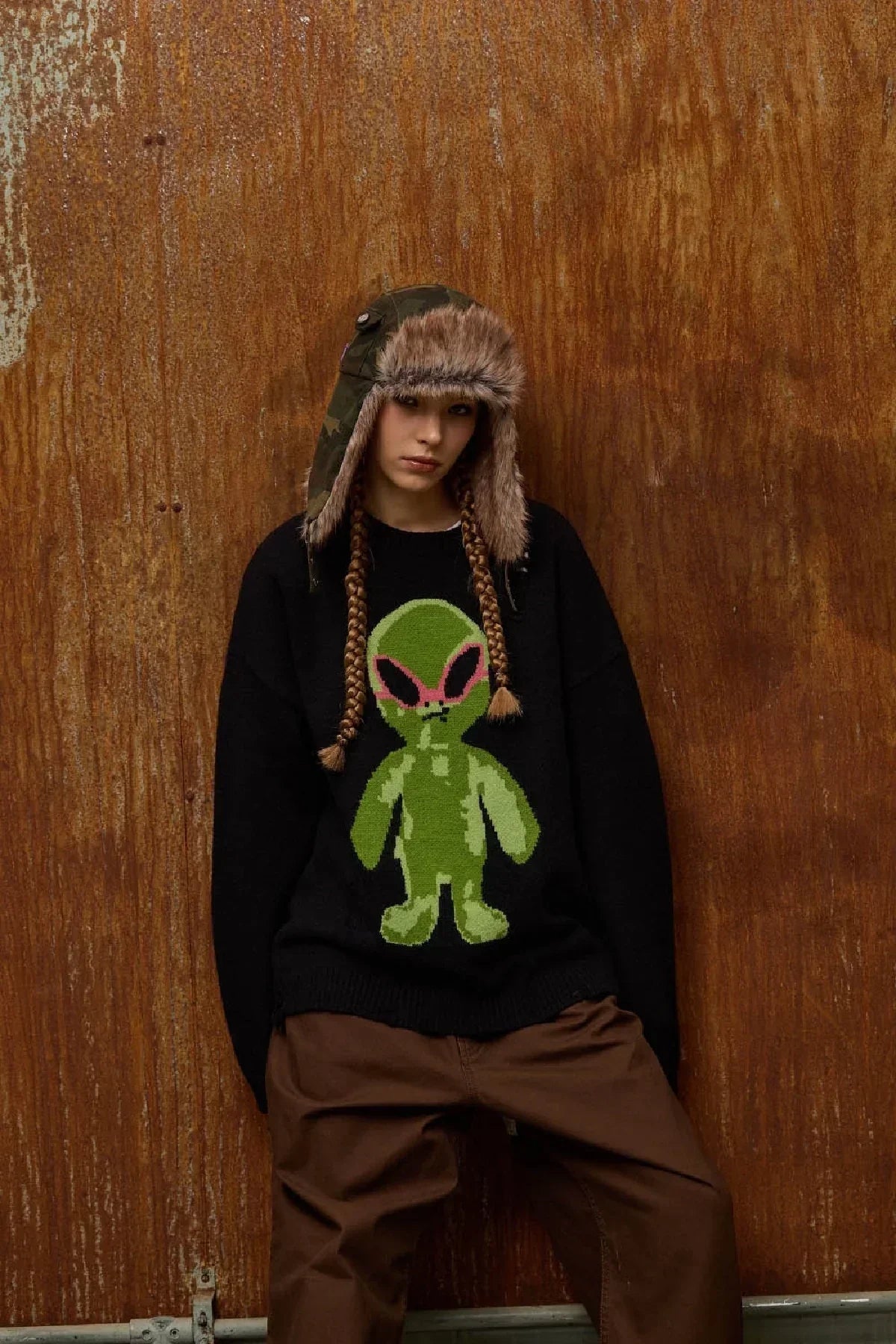 Person wearing camo fur trooper hat and black sweater with green alien design standing against rusty brown wall