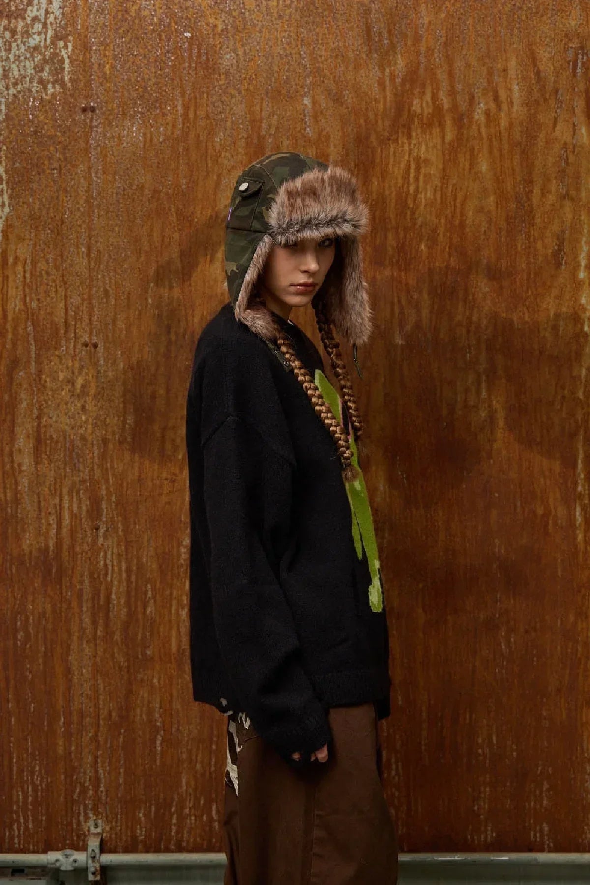 Model wearing camo fur trooper hat, black sweater, and brown pants against rusted metal background
