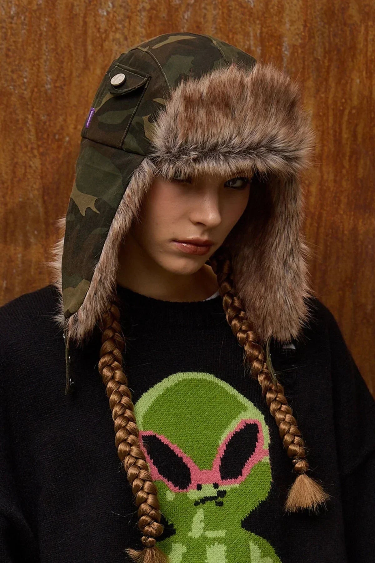 Person wearing camo fur trooper hat and black sweater with green alien graphic and pink sunglasses