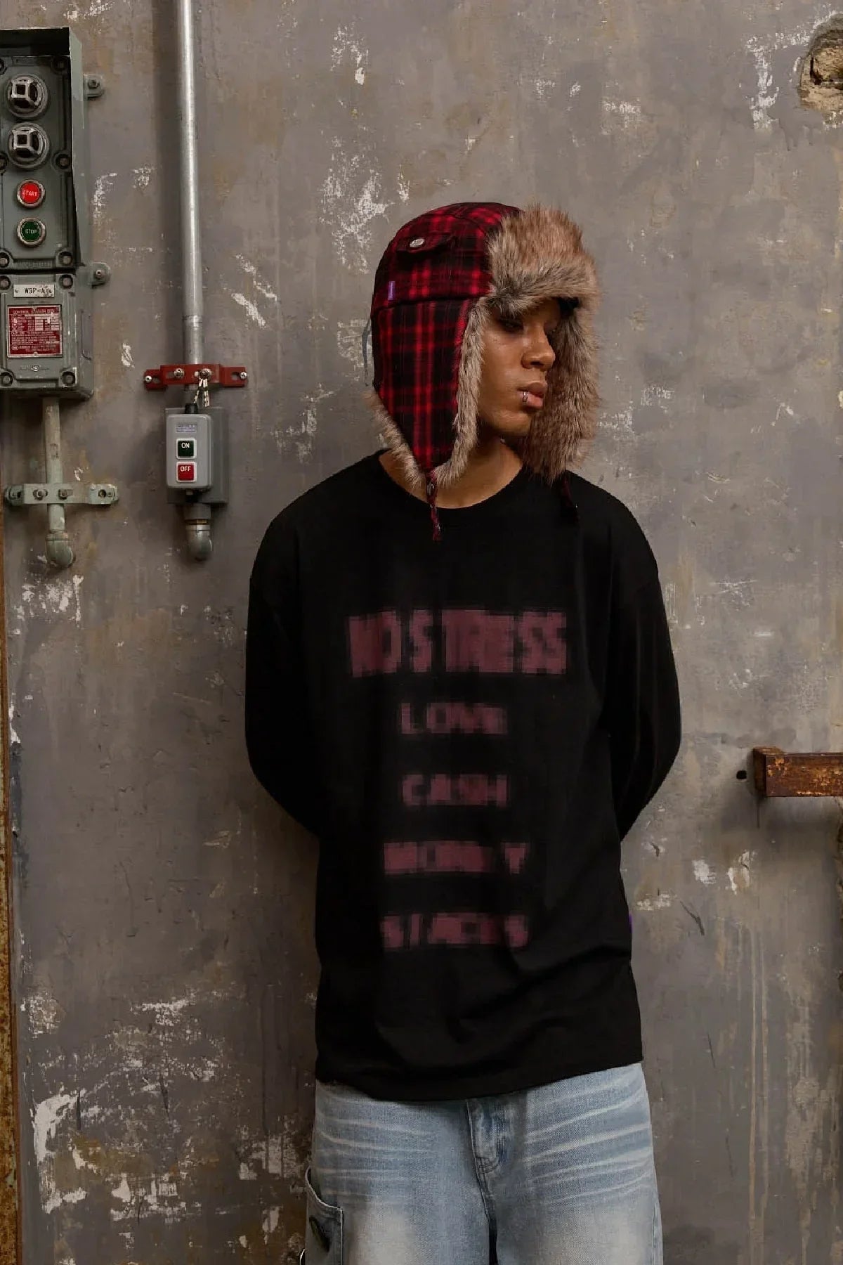 Man wearing red check fur trooper hat and black long sleeve shirt standing against grungy concrete wall