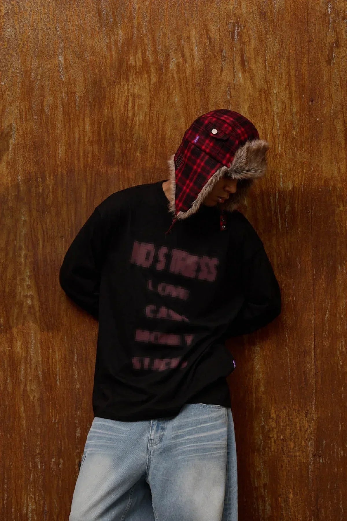 Person wearing red checkered fur trooper hat and black graphic sweatshirt against textured brown wall