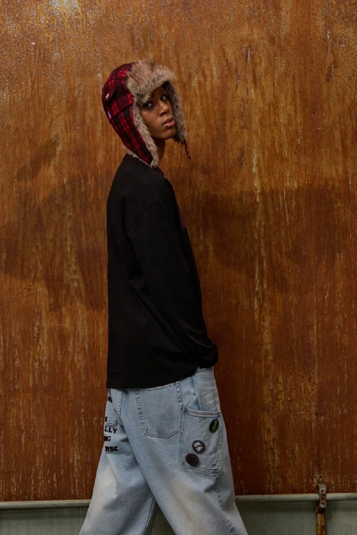 Person wearing red plaid fur trooper hat, black long sleeve shirt, and light blue patched jeans against rusty metal wall