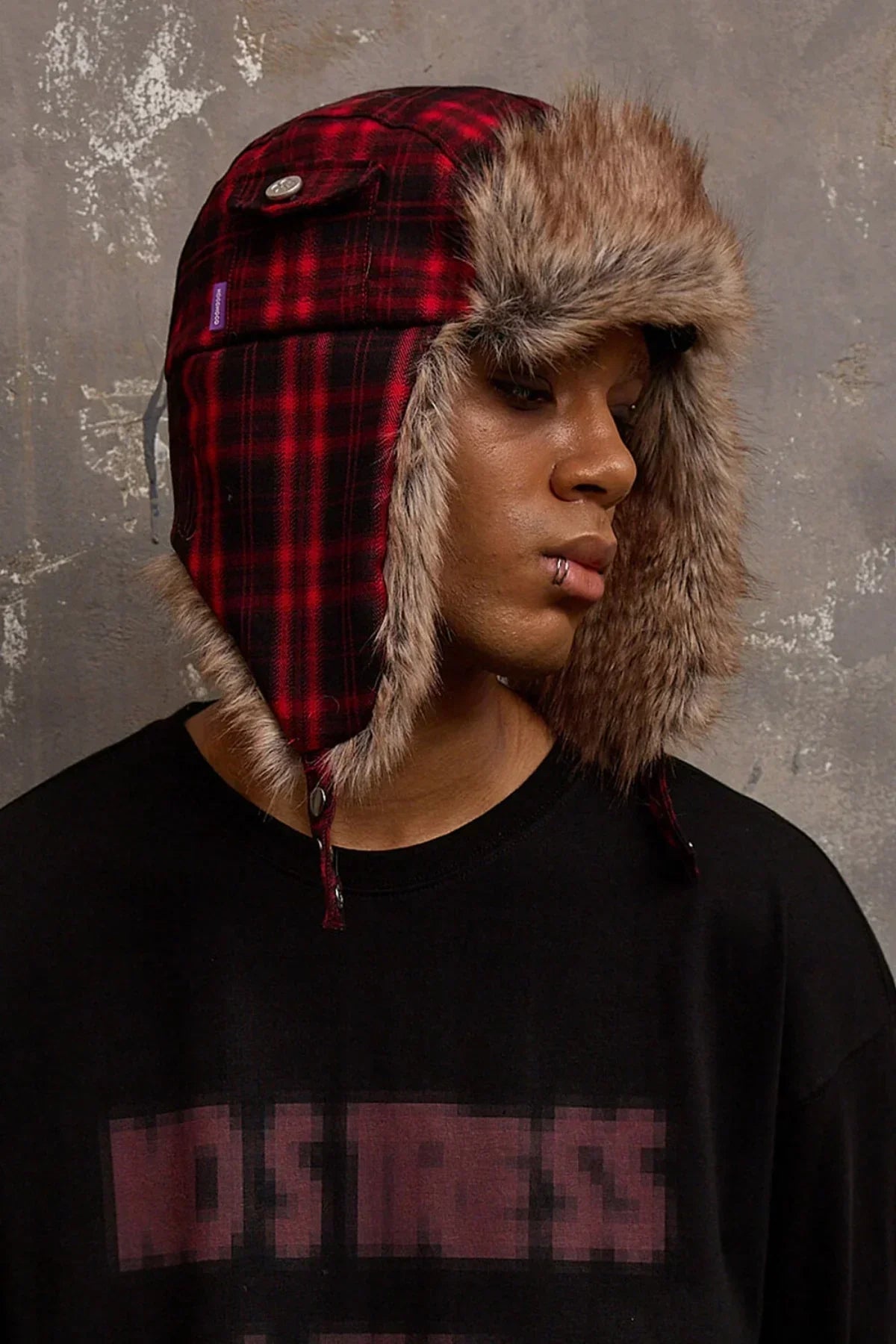 Man wearing red plaid trapper hat with brown faux fur trim and black shirt, against gray textured wall