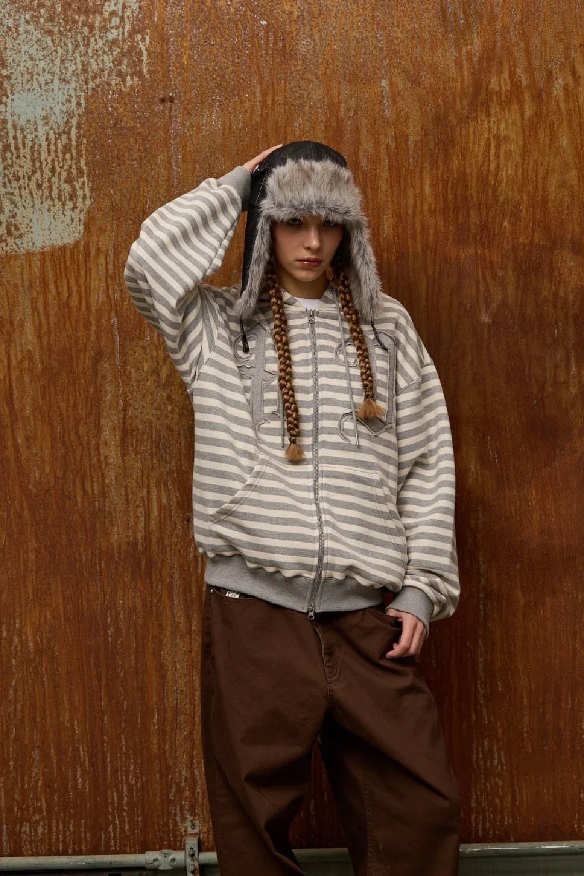 Person wearing black denim fur trooper hat, gray striped zip-up hoodie, and brown pants against rusted metal wall