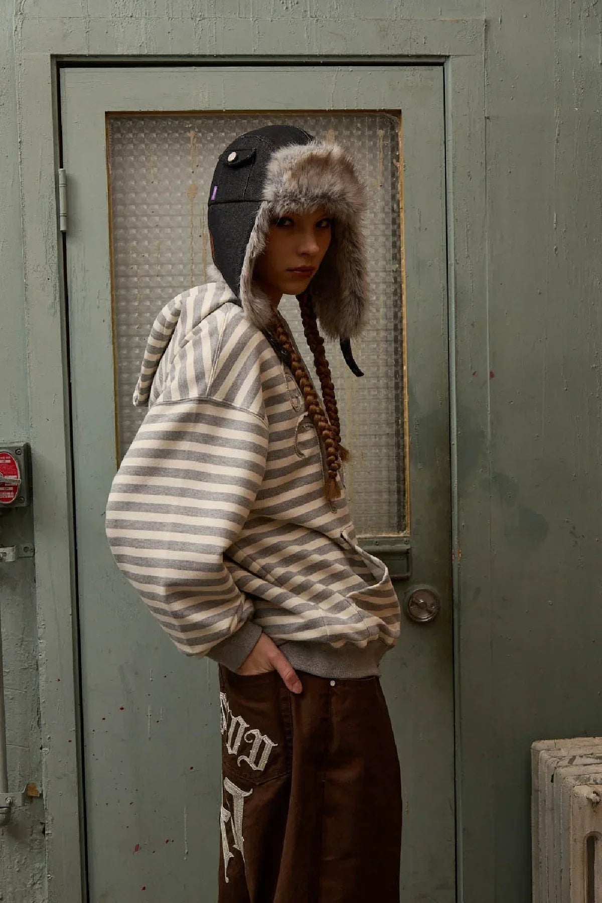 Model wearing black fur-trimmed trapper hat and striped hoodie, standing against green door backdrop