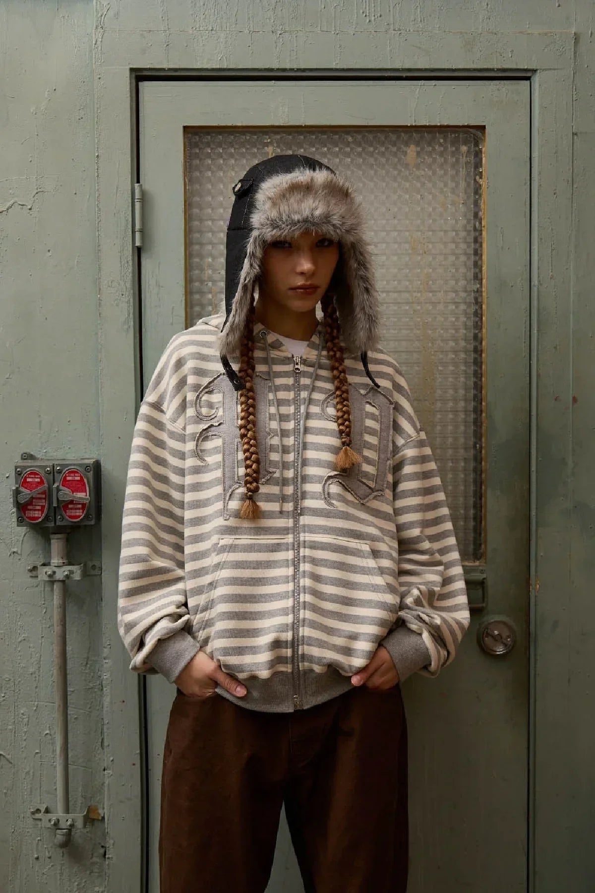 Person wearing a black fur-trimmed trooper hat, striped zip-up hoodie, and brown pants indoors