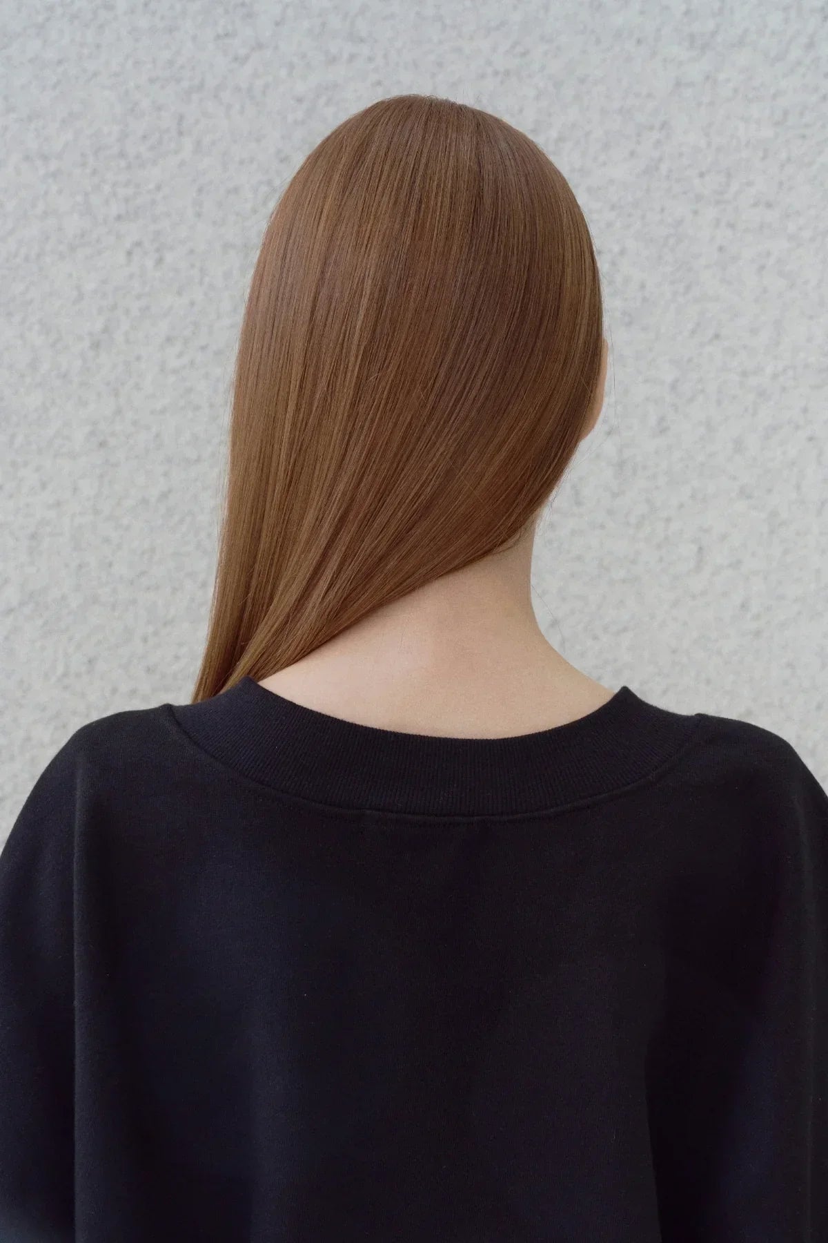 Close-up of a person with long straight brown hair wearing a black heavy cotton sweatshirt against a textured gray wall