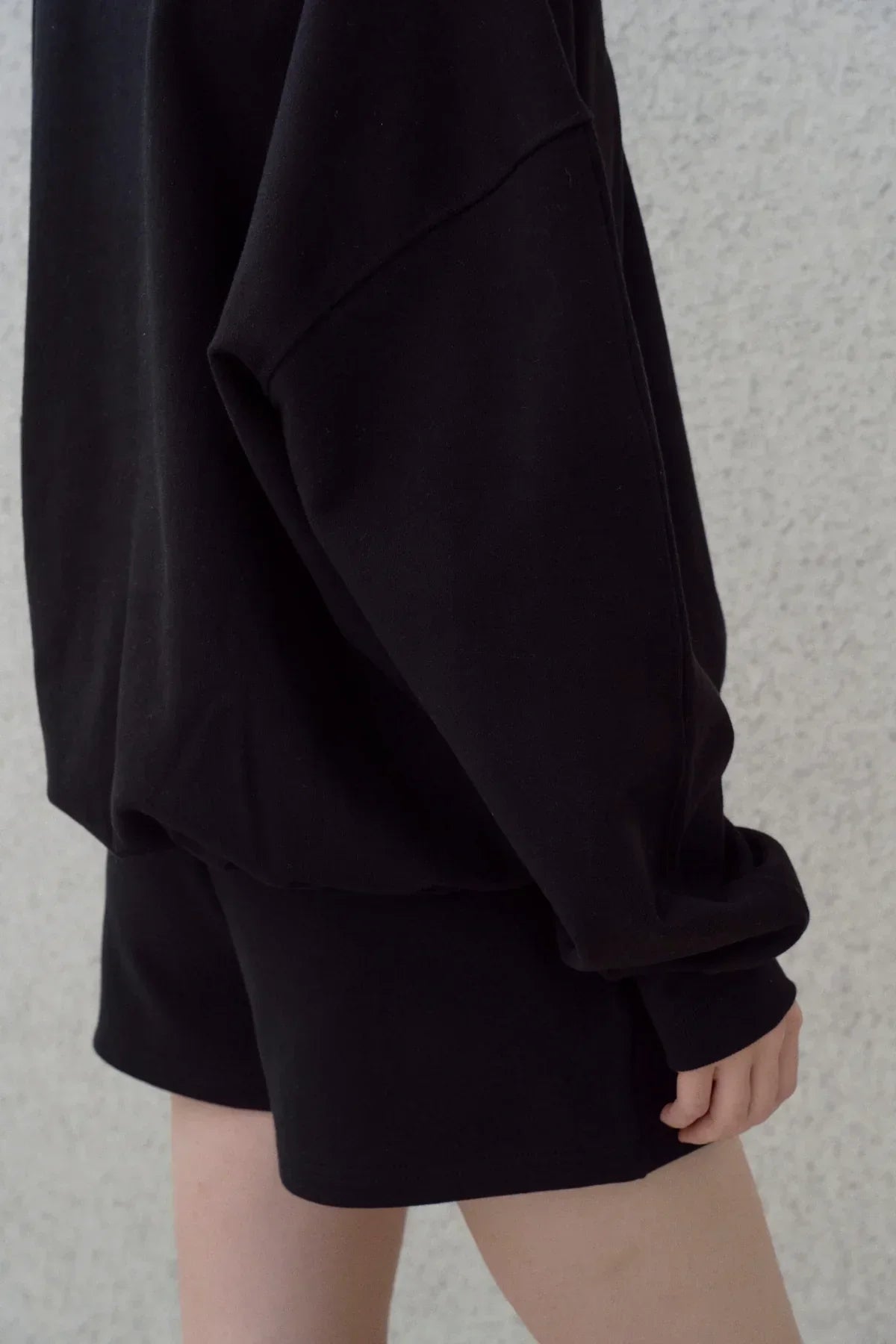 Close-up of black heavy cotton sweatshirt and matching shorts on person against light textured wall
