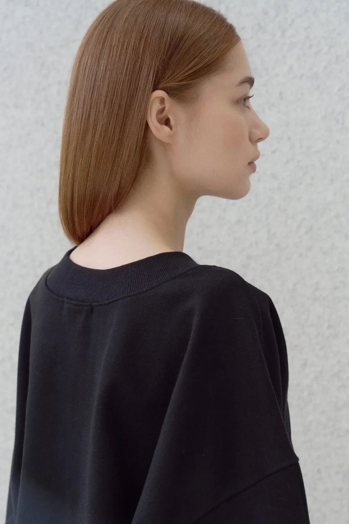 Side profile of woman with straight hair wearing a black heavy cotton sweatshirt against gray wall
