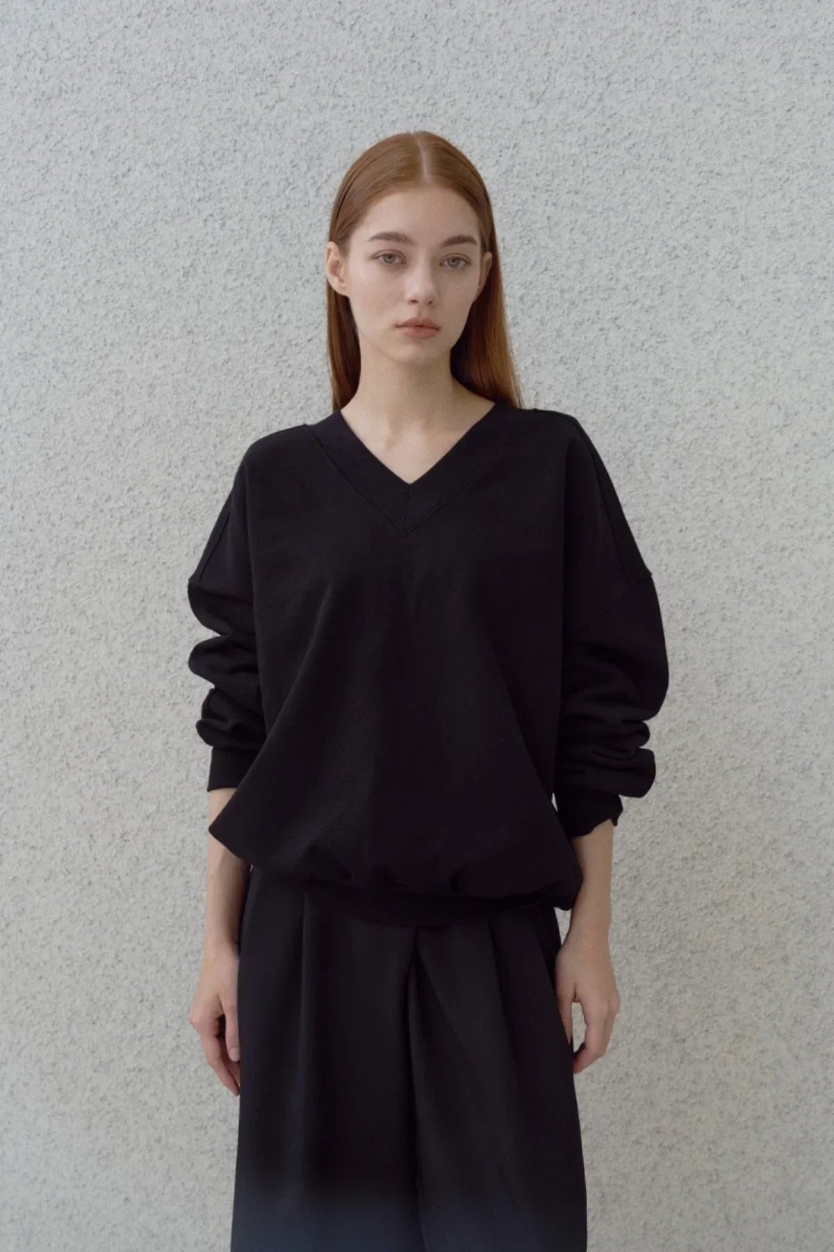Young woman wearing a black heavy cotton V-neck sweatshirt and black pants standing against a gray textured wall