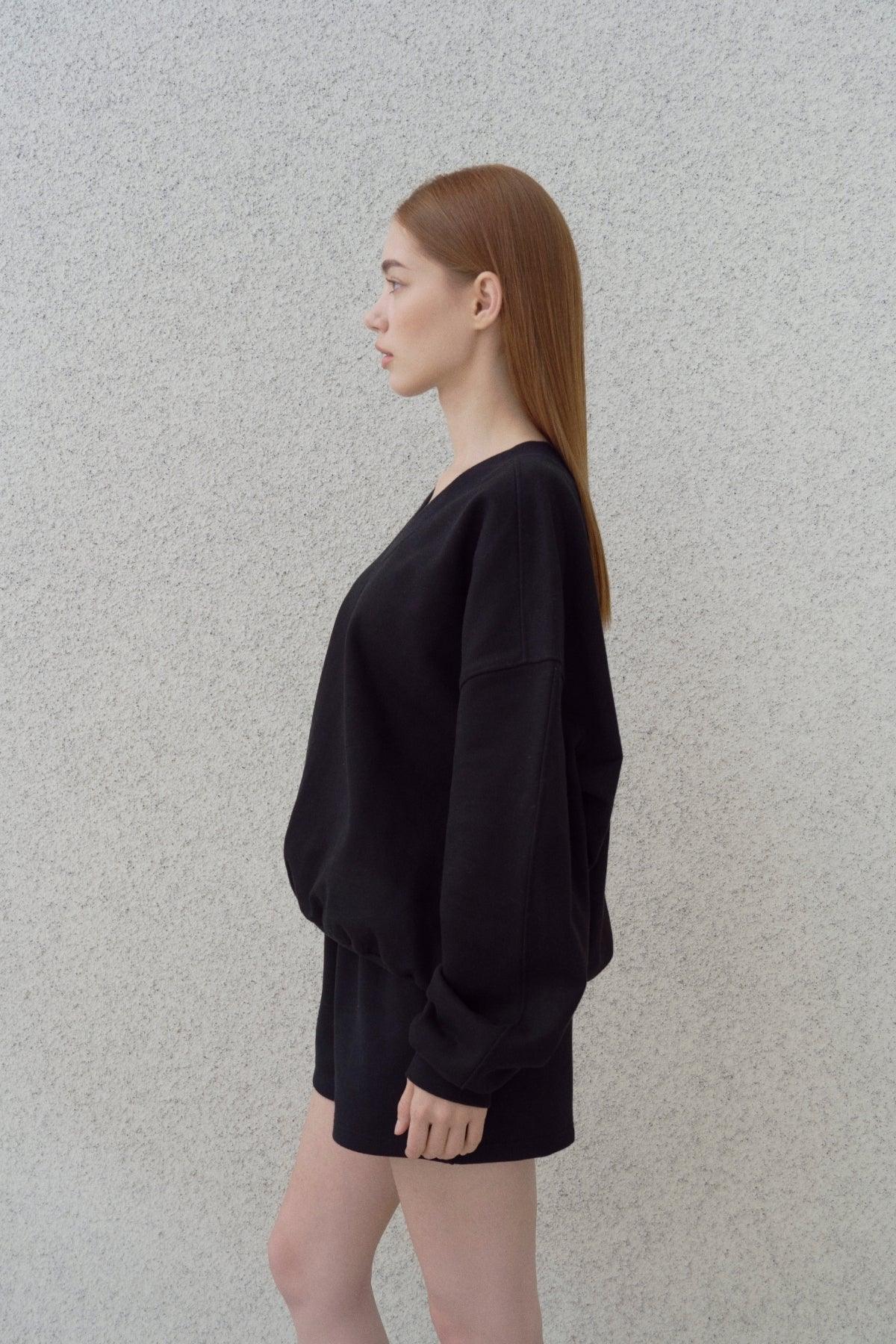 Side view of woman with long red hair wearing black heavy cotton V-neck sweatshirt and shorts against gray background