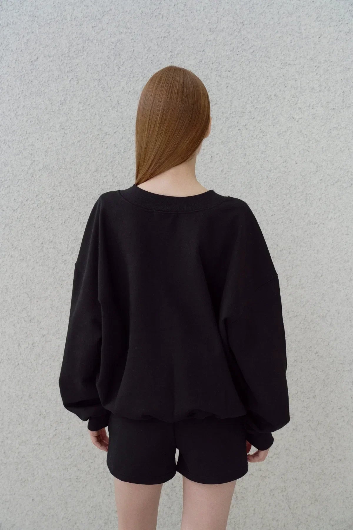 Back view of a person wearing a black oversized sweatshirt and black shorts against a gray textured wall