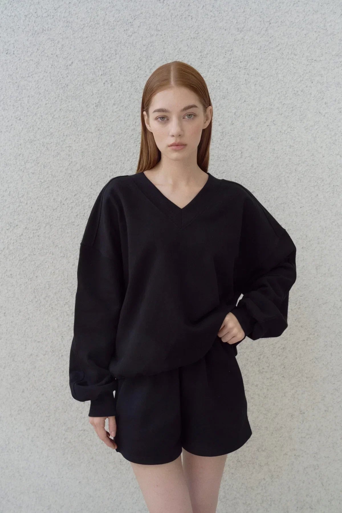 Young woman wearing a black heavy cotton V-neck sweatshirt and matching shorts against a textured gray wall