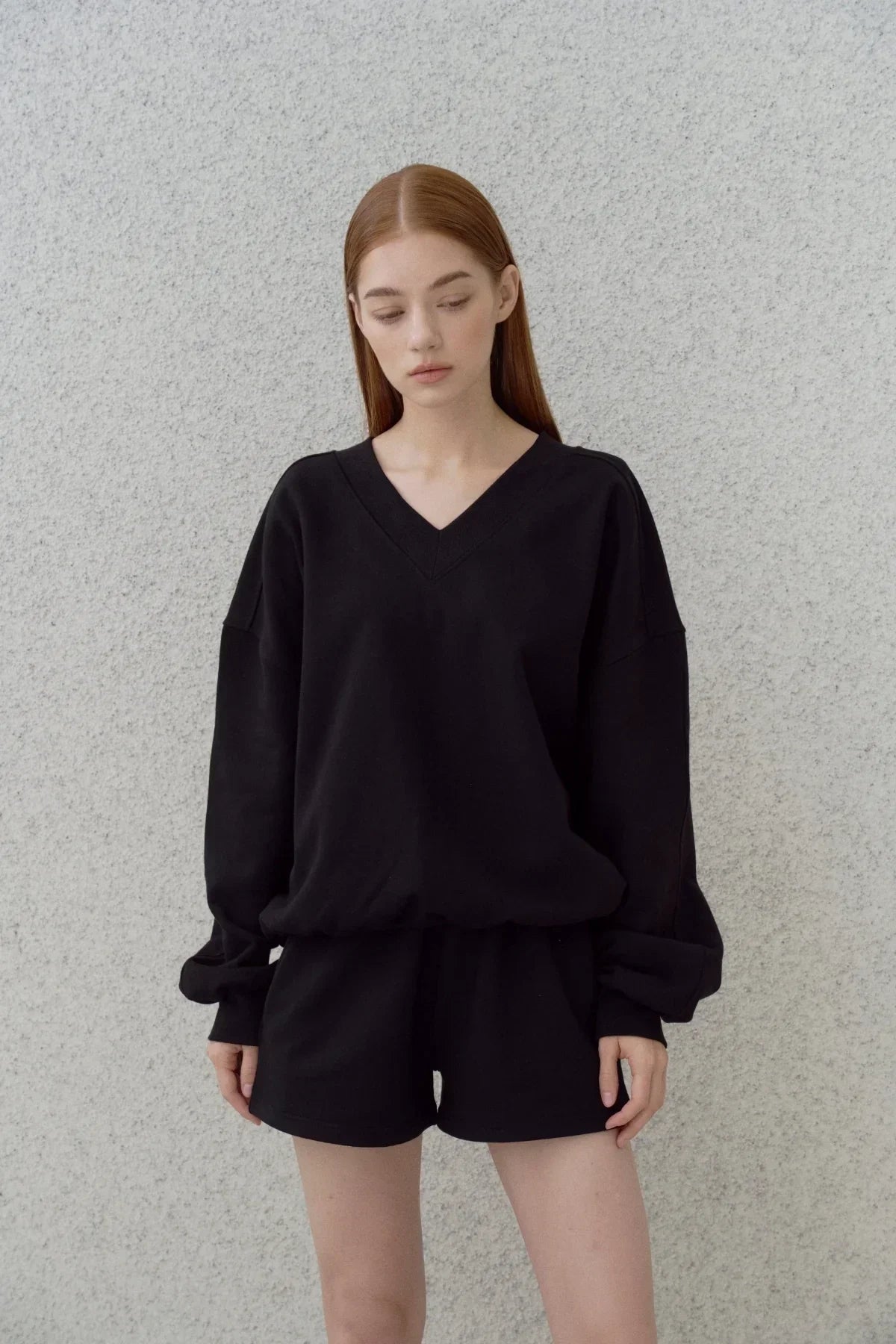 Young woman wearing black heavy cotton V-neck sweatshirt and matching shorts against a textured light gray wall